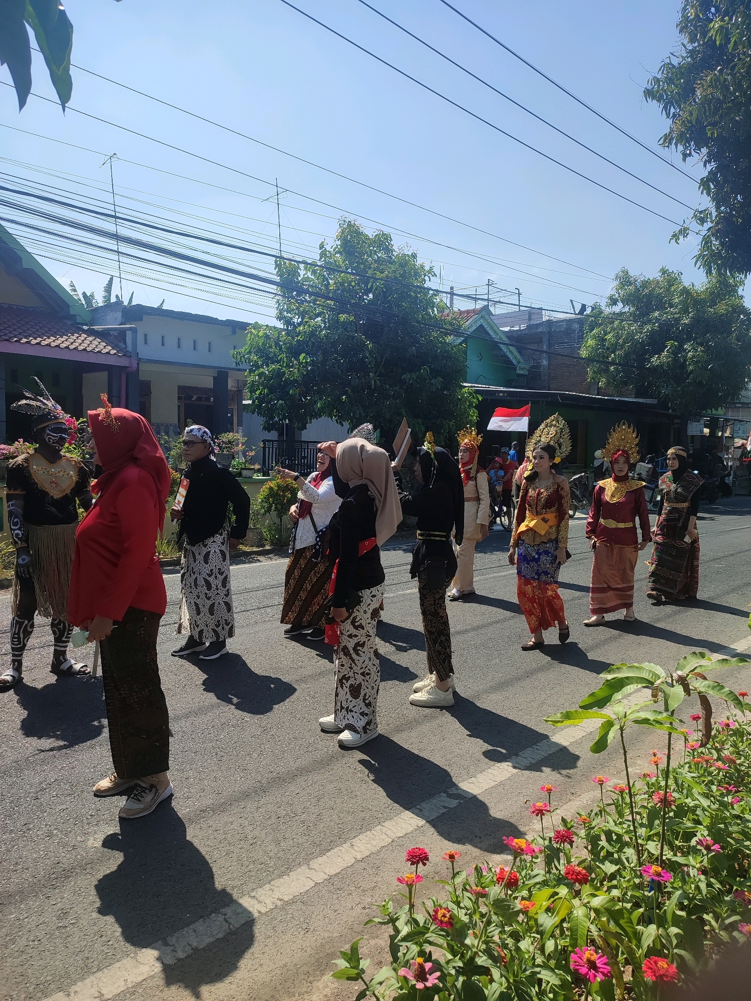 The image depicts a street scene in Nganjuk, Indonesia, with a group of people in traditional attire walking on the asphalt road. The people are diverse in age and gender, and many are adorned in colorful costumes, some with elaborate headwear. Several individuals are wearing batik patterns on their clothing, which is a traditional Indonesian fabric. A few people are seen holding flags, including one prominently displaying the Indonesian flag. The background reveals residential buildings and trees, suggesting a neighborhood setting. The sky is clear and blue, indicating a sunny day. The presence of flowers in the foreground adds a touch of natural beauty to the urban environment. The overall atmosphere appears festive, possibly indicating a parade or cultural event.
