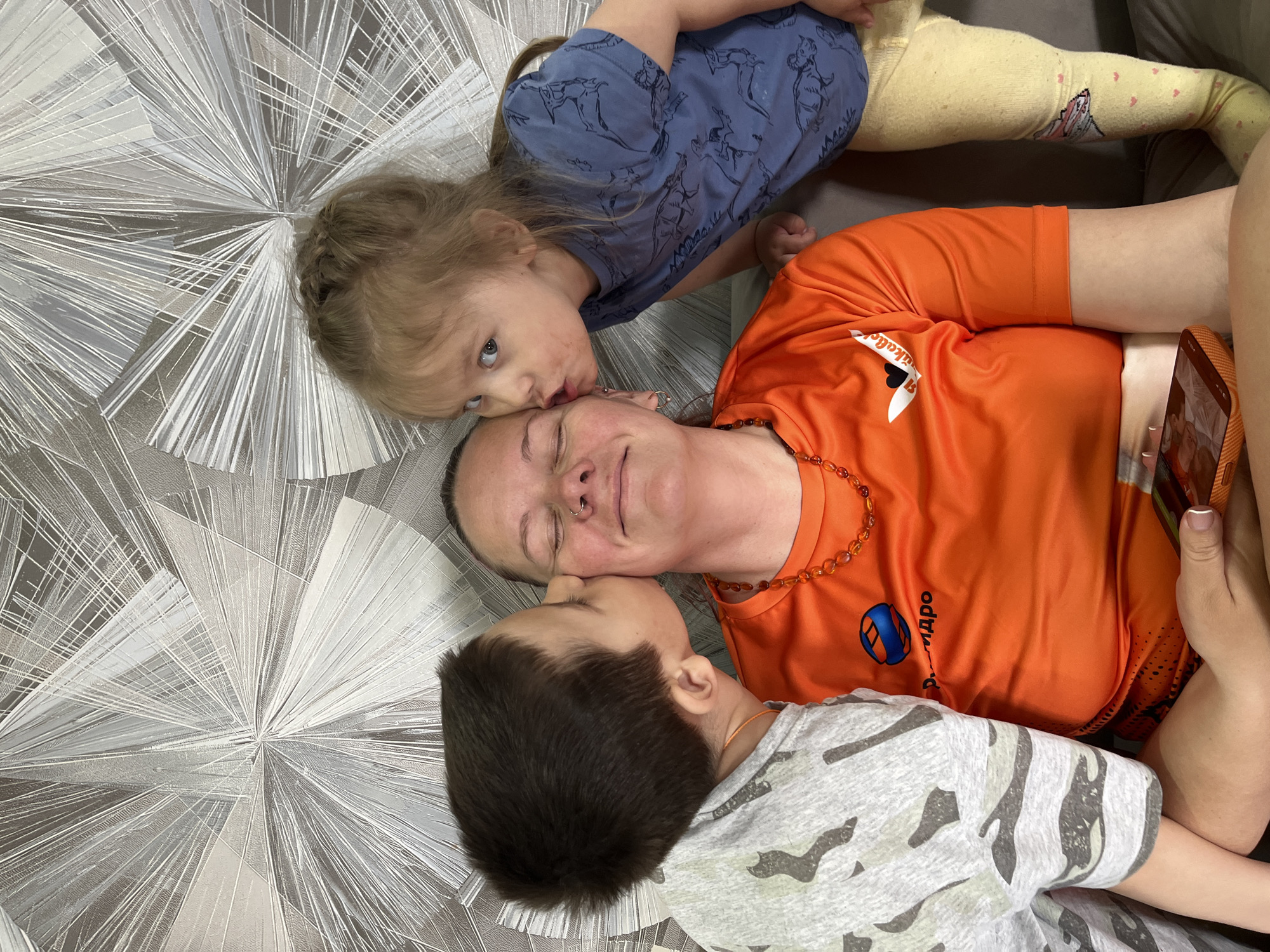 An overhead view captures an intimate family moment indoors in Chaikovsky, Russia, featuring an adult and two young children lying closely together. The adult, likely a woman, lies on her back with her eyes closed in a peaceful expression. She wears a bright orange short-sleeved shirt with the text "Olimpiadro" and a blue and orange volleyball-like logo printed on it. A multi-strand amber-colored beaded necklace is visible around her neck, and she has a nose piercing.
To her upper left, a young girl with blonde hair, dressed in a blue t-shirt with dark animal patterns and light yellow leggings, is shown with her lips pressed against the adult's cheek, appearing to give a kiss. To the adult's lower right, a young boy with short dark hair, wearing a grey camouflage-patterned t-shirt, also leans in with his face near the adult's cheek, indicating a similar gesture of affection.
The adult holds a smartphone in her right hand, partially visible, displaying images of people on its screen. They are all positioned on a distinctively patterned surface, which could be a rug, blanket, or wallpaper. This background features an intricate, radial design of light grey and white lines emanating from multiple points, creating a dynamic, abstract visual effect. The overall scene conveys warmth, affection, and a relaxed family atmosphere.