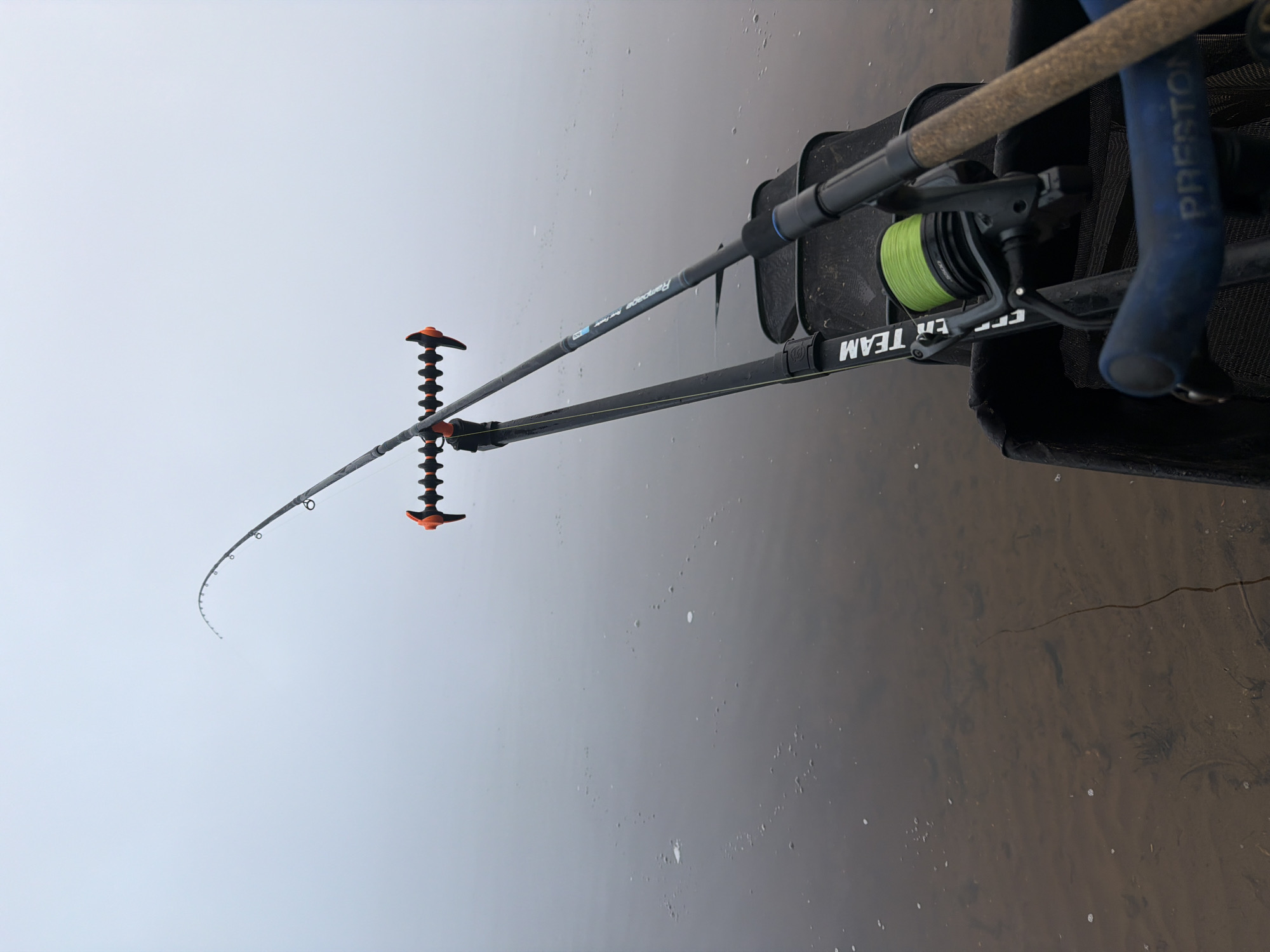 The image, set in Noviy, Russia, displays a fishing setup by the edge of a body of water under an overcast sky. Two fishing rods are prominent in the frame, set up as if actively fishing. The main rod, a "Rampage Feeder" model, extends over the water and is supported by an orange and black "snake-style" rod rest. A black fishing reel with a vibrant green line is attached to this rod, showing "DAIWA" branding and "4000" as a model number. Behind it, a second rod is partially visible, with "Preston" inscribed on its blue handle. Both rods are positioned next to a dark bag or container, possibly a keepnet or tackle bag, which features the text "FER TEAM" (likely "FEEDER TEAM") on its strap.
The sky is a uniform light grey, indicating heavily overcast weather. The water appears shallow near the bank, revealing a muddy or sandy bottom with subtle textures and a few small bubbles on the surface. The scene suggests a calm fishing session in progress.