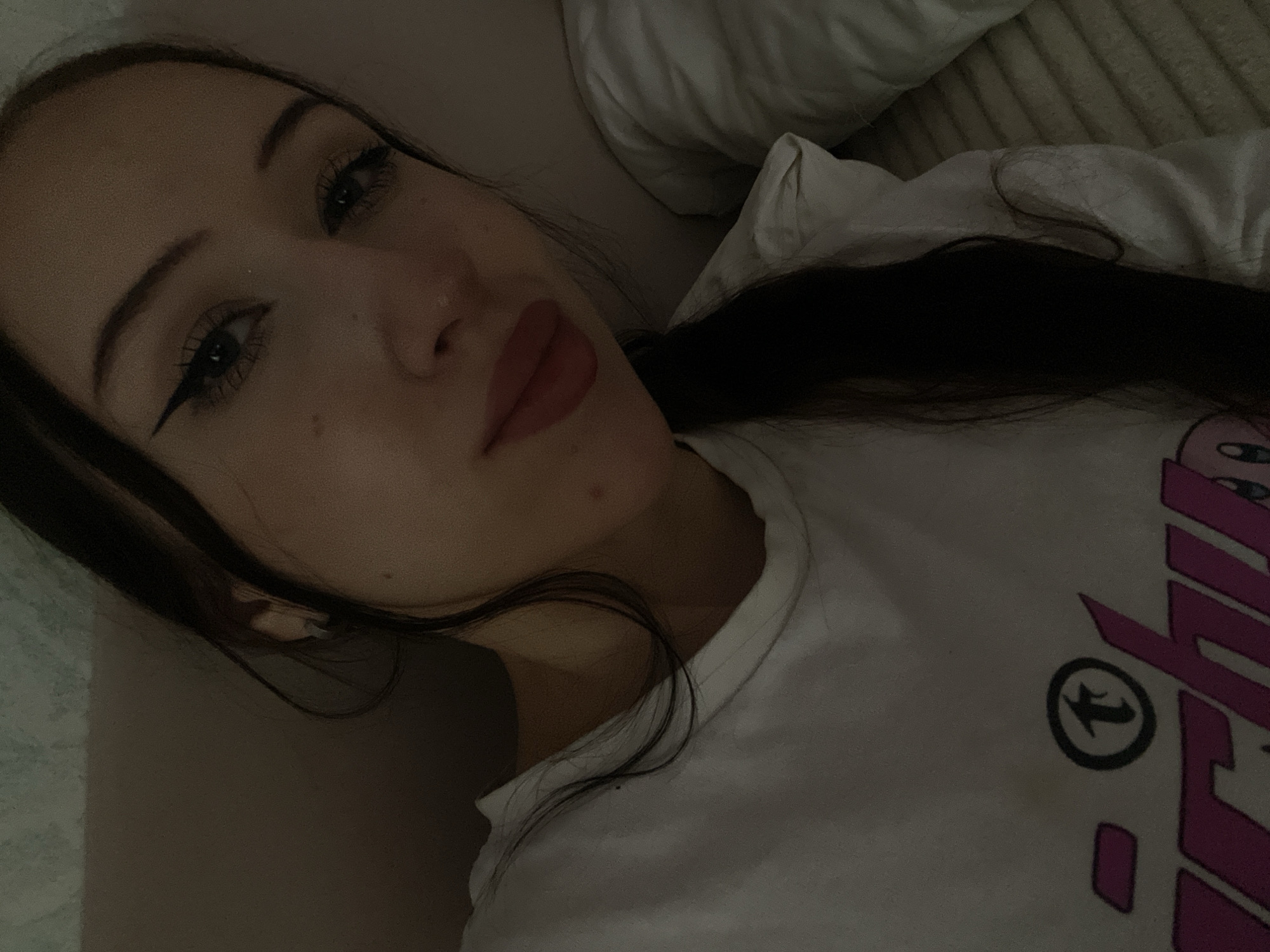The image is a close-up selfie of a young woman with long, dark hair, lying down on what appears to be a bed with a striped pillow. She is wearing a white t-shirt with a graphic logo. The lighting is dim, creating a soft, intimate atmosphere. Her makeup includes winged eyeliner and lip gloss. The background is a textured wall. The image does not contain any information about the city of Votkinsk, Russia.