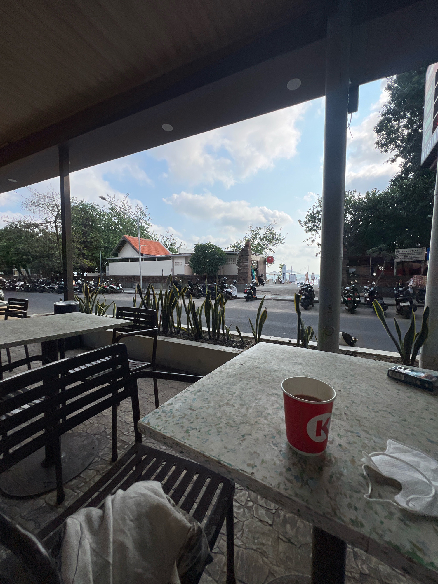 This is a medium shot of an outdoor seating area at a cafe in Kuta, Indonesia. In the foreground, a speckled grey table is set with a red paper cup of dark liquid, a disposable mask, and a pack of cigarettes. A white cloth is draped over a black metal chair to the left. Beyond the table, a street is visible, lined with parked motorcycles and trees. Buildings with orange roofs and white walls are situated across the street. In the background, the sky is partly cloudy. A sign with Indonesian text is visible on a building to the right.