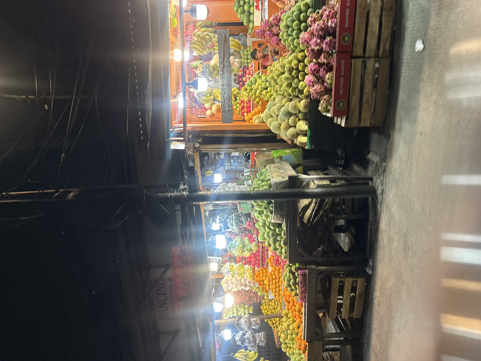 The image depicts a fruit stand in North Jakarta, Indonesia. The stand is brightly lit with numerous light bulbs, showcasing a colorful array of fruits including bananas, mangoes, oranges, dragon fruits, and various other tropical fruits. The fruits are stacked in wooden crates and on tiered stands. A person is visible in the background. Power lines and cables crisscross above the stand. A sign with the word "PANCING" is seen in the background. The ground appears to be concrete. The surrounding environment is dark, suggesting the photo was taken at night.