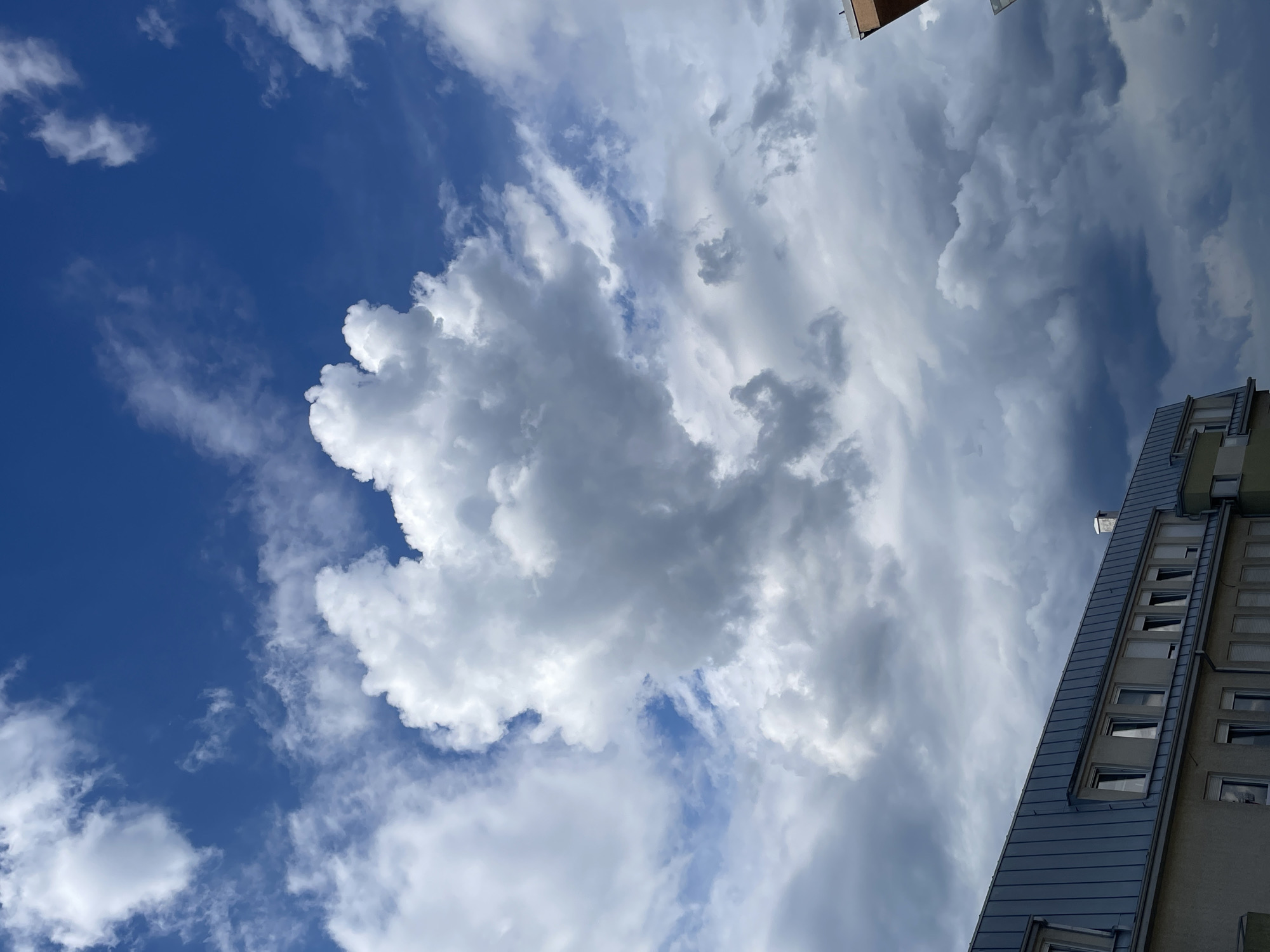 The image depicts a vibrant, partly cloudy sky over a residential or commercial building in the city of Unknown, Unknown. The upper two-thirds of the frame are dominated by dynamic cloud formations against a deep blue sky. Large, bright white cumulus clouds are prominently featured, with a central, substantial cloud exhibiting a somewhat bulbous, almost heart-like shape. Towards the bottom and right of the sky, the clouds transition to darker, denser gray masses, suggesting an approaching weather front or localized overcast conditions. Patches of clear blue sky are visible in the upper left and right, and between the white cloud formations.
The lower third of the image features the upper section of a multi-story building. It has a distinctive light gray, possibly metallic, mansard-style roof with a series of integrated rectangular dormer windows, some open and reflecting the sky. Below the roofline, a facade in a light beige or pale green hue is visible, featuring a row of rectangular windows. Many of these windows are equipped with closed white roller shutters, while others are open or partially open. A small, metallic chimney stack is visible on the right side of the main roof. On the far right edge of the frame, a partial corner of an adjacent building with an orange-brown upper section is visible. No individuals or specific actions are discernible within the frame.