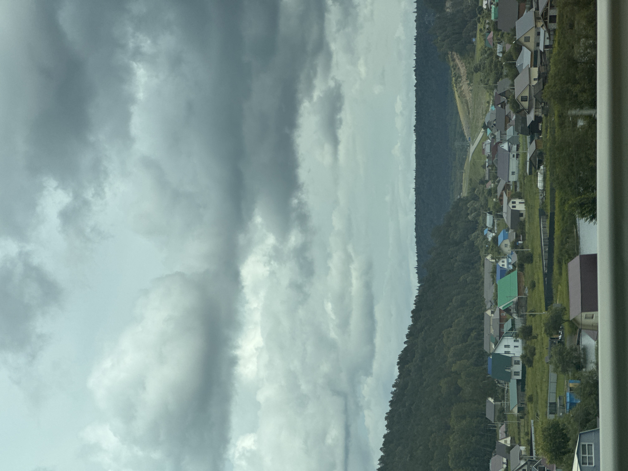 The image captures a rural settlement in Baiki, Russia, characterized by a cluster of houses nestled amidst rolling hills and dense forest. The sky is overcast with dramatic, dark clouds, creating a somber atmosphere. In the foreground, a body of water is visible, reflecting the muted light. The houses are predominantly single-story structures with pitched roofs in various colors, including green, grey, and blue. Some appear to be made of wood, while others have more modern finishes. The landscape is verdant, with trees covering the hillsides and scattered around the village. A road winds through the scene, leading towards a distant horizon. The overall impression is one of a peaceful, remote community existing in harmony with its natural surroundings.