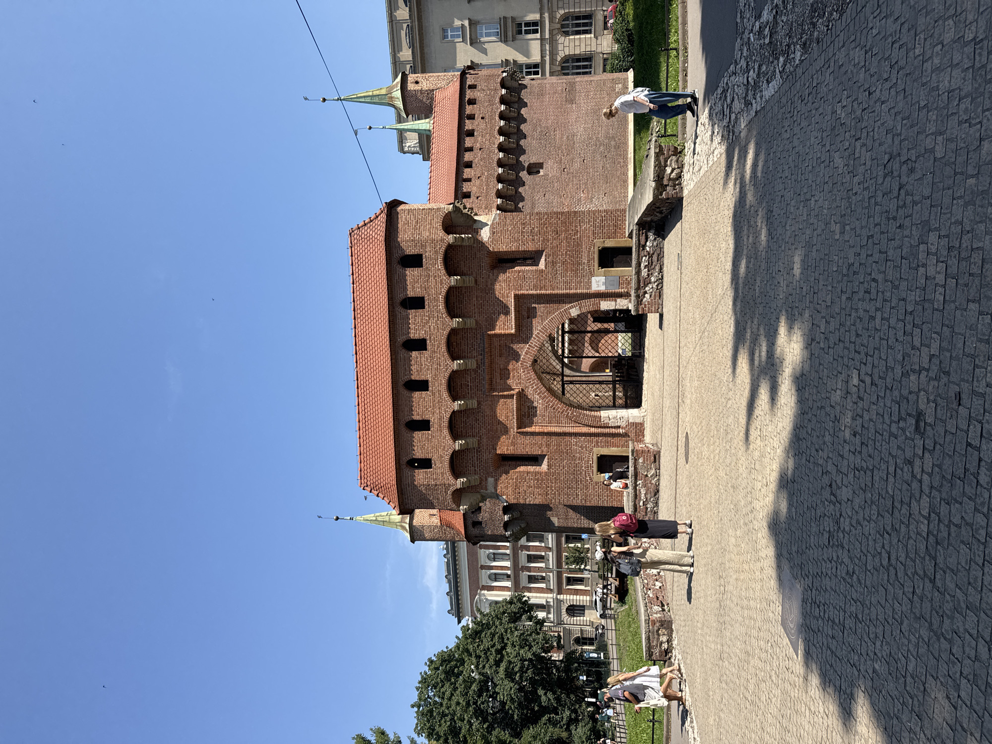 The image depicts the Krakow Barbican in Krakow, Poland, on a clear, sunny day. The Barbican, a medieval fortified outpost, is constructed from red brick and features a prominent arched entryway. Two green-topped towers rise from the structure, adding to its historic appearance. The foreground is paved with cobblestones, with dappled sunlight creating patterns on the ground. Several people are visible, including a woman in a white dress and a man walking together on the left, and two women standing in the center, one wearing a red backpack. On the right, a man in a white shirt and blue jeans walks along a sidewalk. Behind the Barbican, older buildings and trees are visible, contributing to the urban landscape.