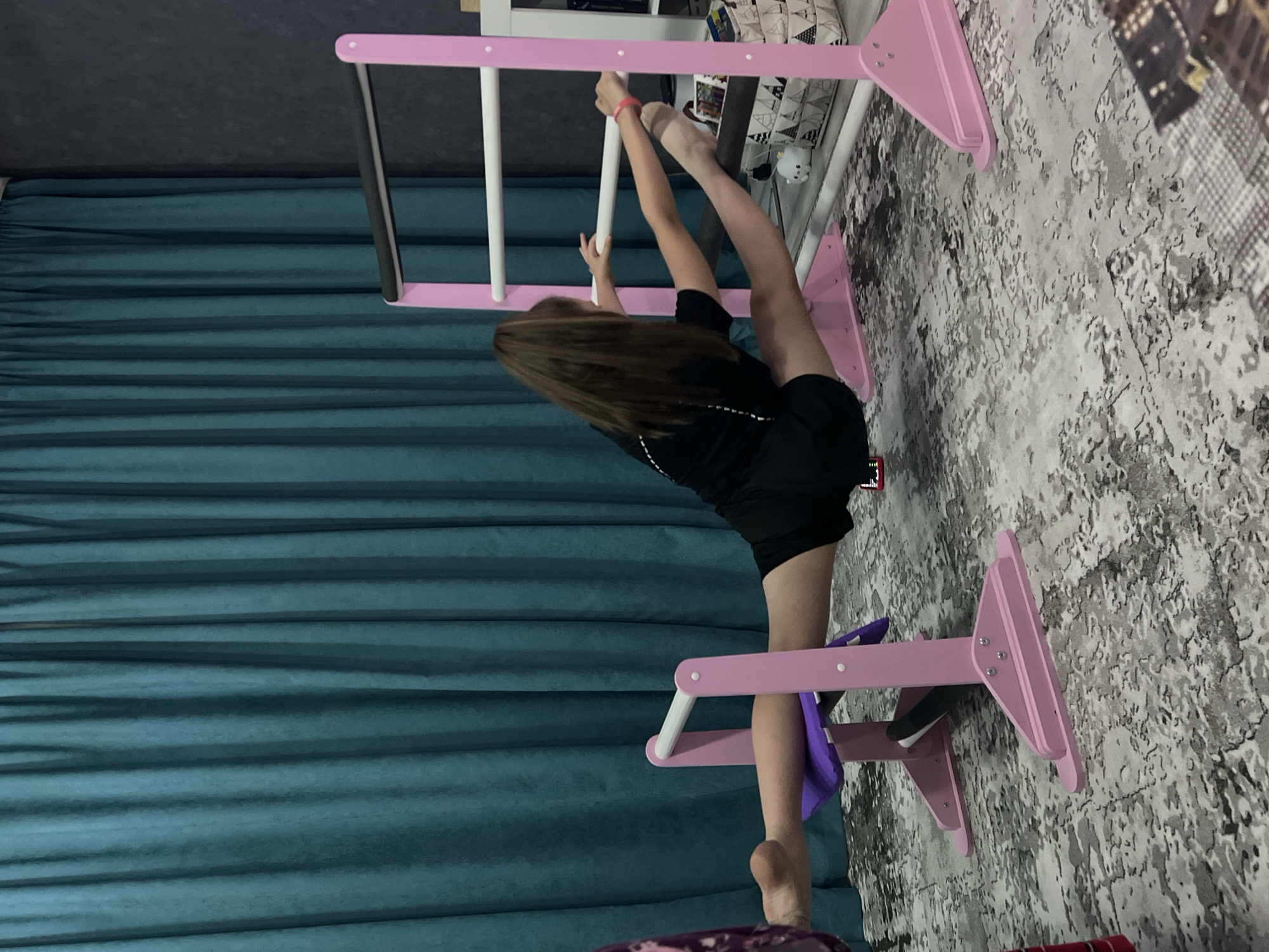 The image shows a person, presumably a child or adolescent, practicing a split on a pink and white gymnastic bar structure indoors. The person is wearing black clothing. The background includes a teal-colored curtain and a textured gray and white rug or floor covering. Some children's items are seen above the gymnastic bar including, stuffed animals and toys. The location is Chaikovsky, Russia. The person is actively stretching their legs into a split position, using the gymnastic bar for support and balance.