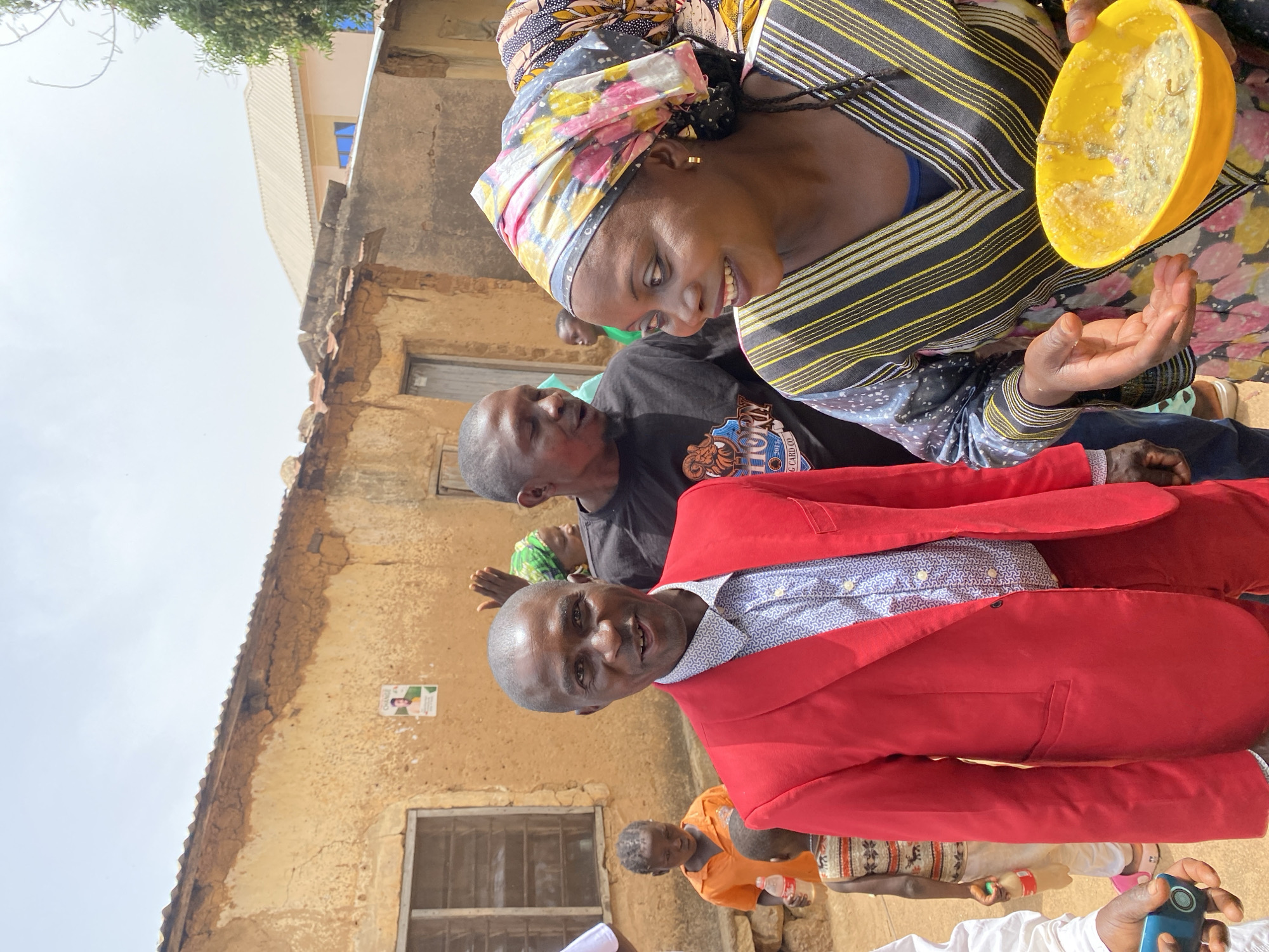 The image depicts a scene in Vom, Nigeria. A woman in a colorful headscarf and striped top holds a yellow bowl, possibly containing food, and extends her hand towards it. A man in a red blazer and patterned shirt stands in front of a building with a window and a poster on the wall. Other people are visible in the background. The setting appears to be outdoors, possibly a gathering or event.