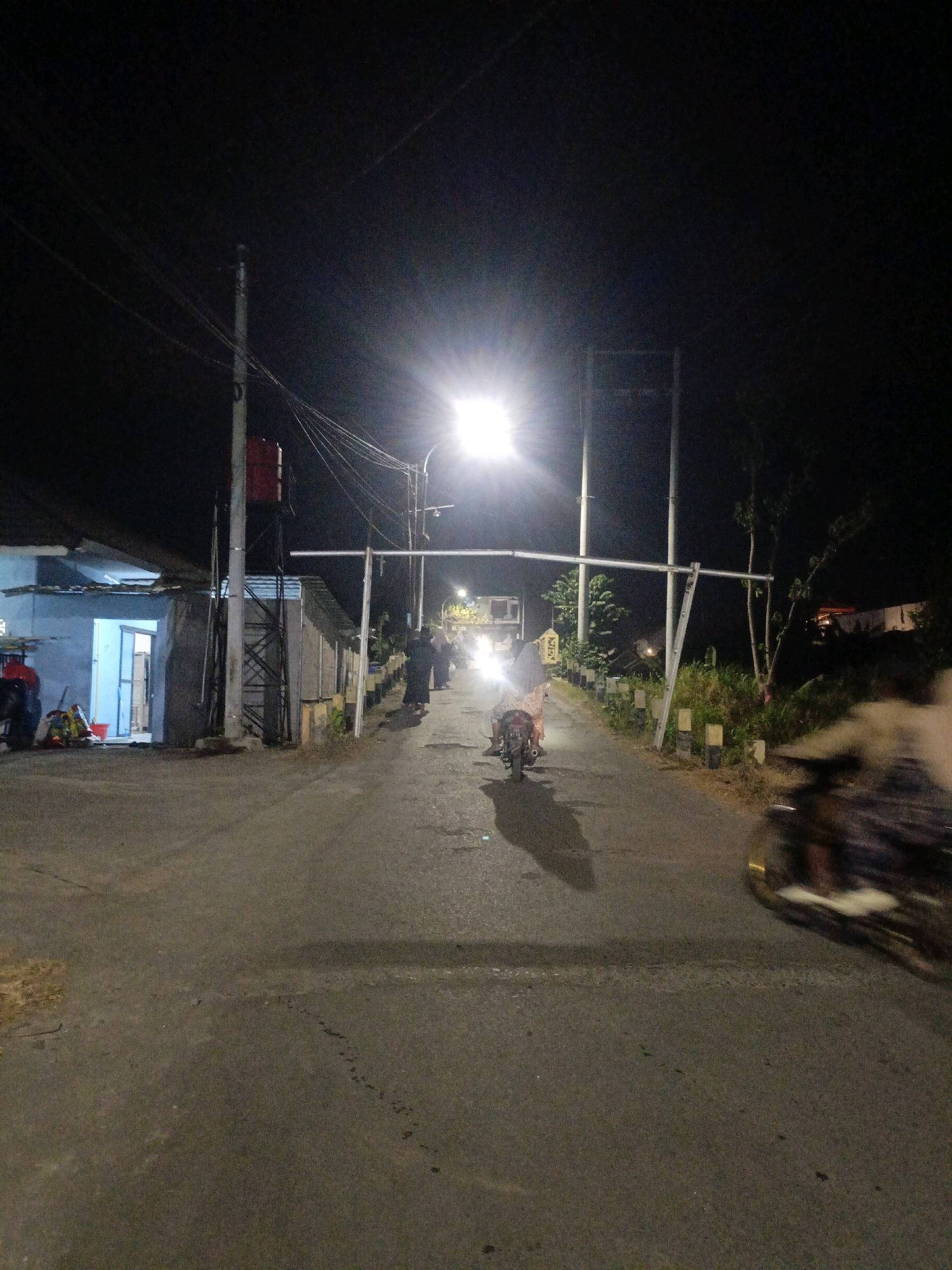 This is a nighttime photograph taken on a paved road in Temenggungan, Indonesia. A bright street light illuminates the scene, casting long shadows. Several people are visible on the road. In the foreground, a person on a motorcycle is captured in motion blur, their body and motorcycle indistinct. Further down the road, another motorcycle with a rider is clearly visible, moving away from the camera. A group of individuals, some appearing to be wearing traditional Islamic attire, are walking on the side of the road, towards the distance. On the left side of the road, a building with blue walls and an open doorway is present, with what appears to be a water tank structure next to it. The environment consists of a road bordered by vegetation and some simple structures. The overall scene depicts a common nighttime street in a residential or semi-rural area.