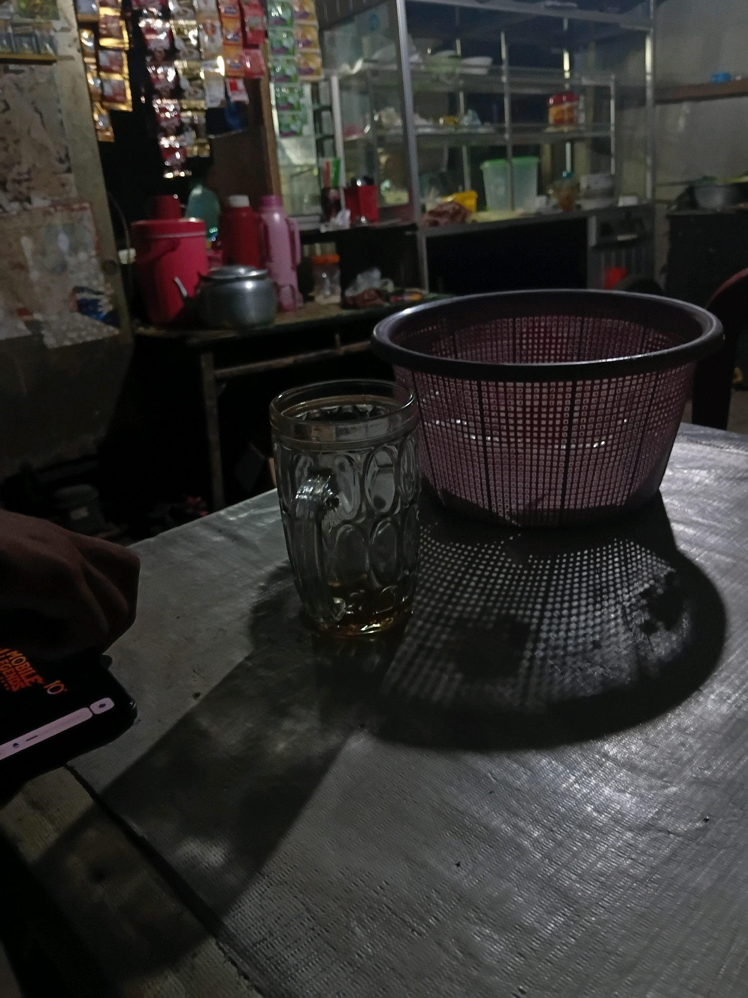 In a dimly lit, possibly outdoor, setting in Bantarbarang, Indonesia, a textured, dark-colored table occupies the foreground. To the right, a red plastic basket with a grid pattern casts a distinct, perforated shadow onto the table. To the left of the basket, a glass mug with a patterned design contains a small amount of amber liquid and is also casting a shadow. A human hand, partially visible on the far left, rests on a dark rectangular object with illuminated text, likely a smartphone displaying "MOBILE LEGENDS". In the background, shelves are visible, stocked with various items including packaged goods, plastic containers, and bottles. Further in the background, more shelves with items are dimly lit, and a glimpse of a refrigerator is visible. The overall impression is of a casual, possibly commercial or domestic, area after dark.