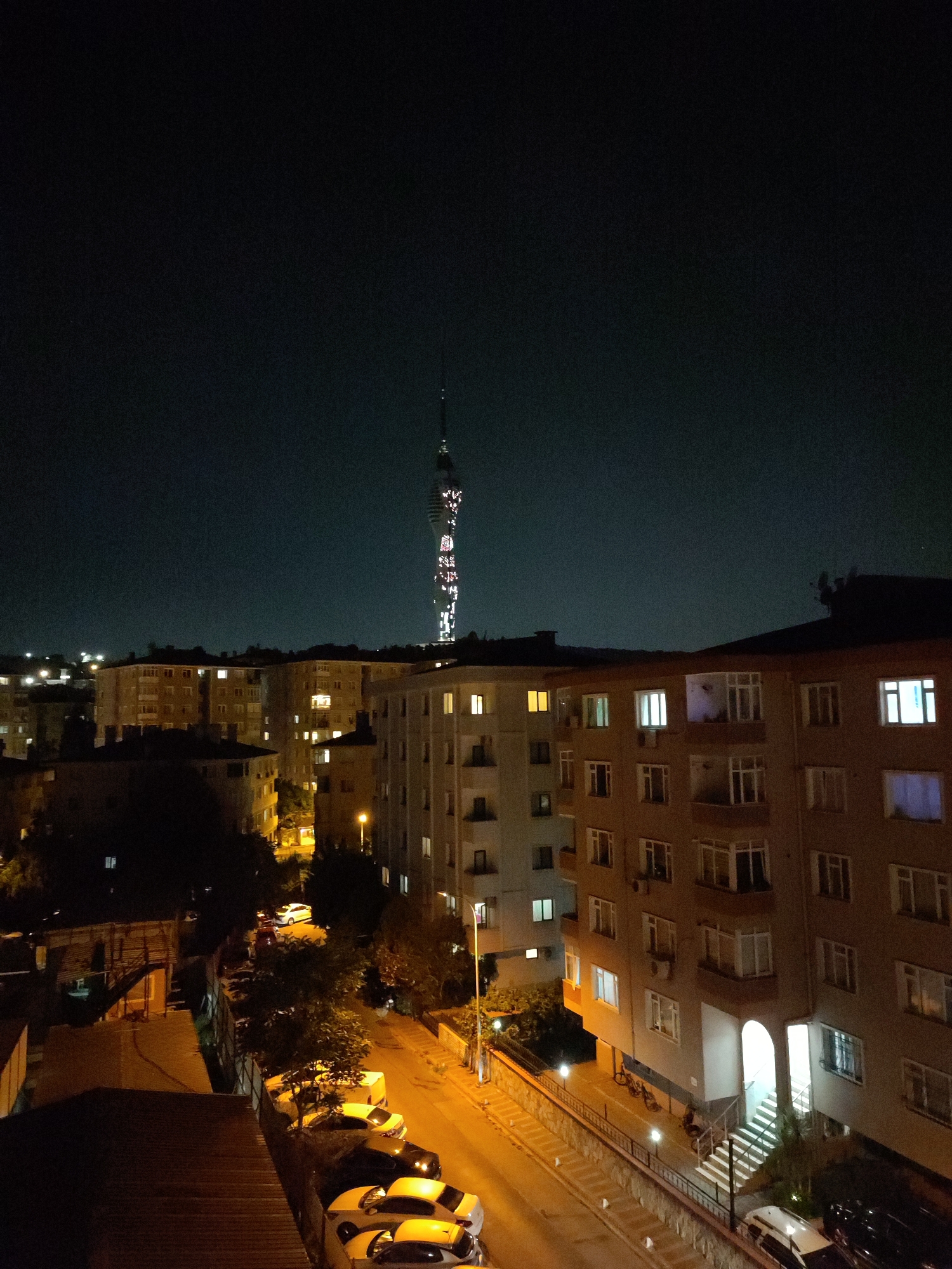 An aerial night view captures the Üsküdar district of Istanbul, Turkey, dominated by the Atakule Tower which stands illuminated against the dark sky. The tower, a prominent landmark, displays a pattern of lights in white and red. Below, a dense urban landscape unfolds with apartment buildings, their windows lit, indicating occupancy. The buildings vary in height and architectural style, with some featuring balconies and exposed staircases. Streetlights cast a warm glow on a road below, where several cars are parked along the side. The overall impression is one of a bustling city at night, with the illuminated tower serving as a focal point in the distance.