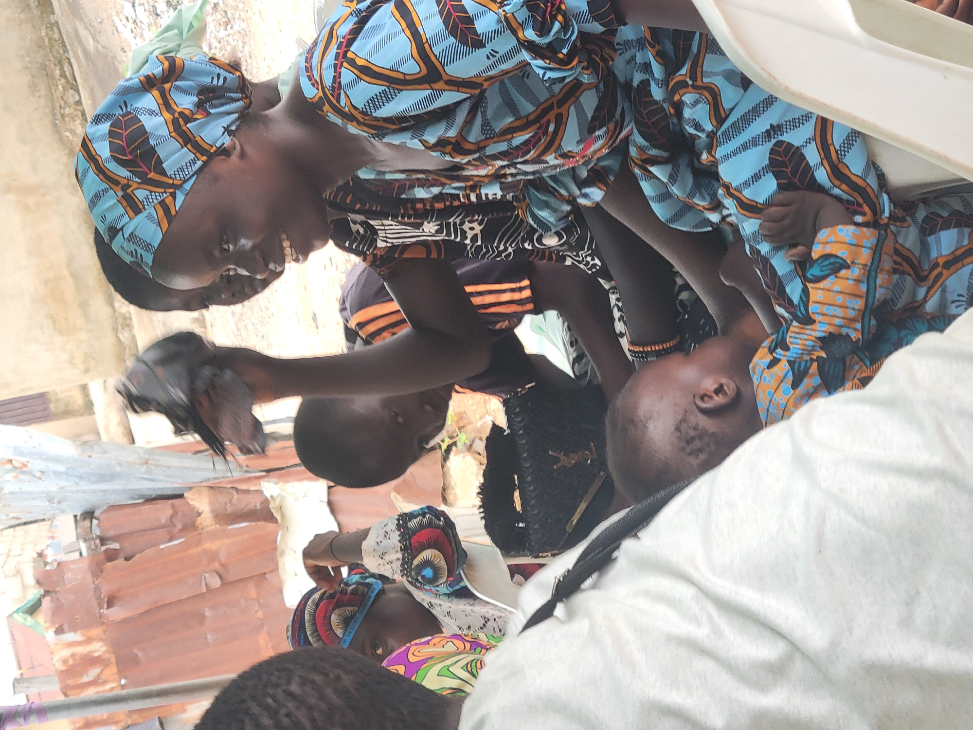 In Madalla, Nigeria, a group of people are gathered outdoors. A woman wearing a blue and orange patterned headwrap and matching dress is holding a dark-colored bird, possibly a chicken or pigeon, in her left hand. She is smiling. Other people are standing around, including children and adults, some wearing patterned head coverings and clothing. The background includes corrugated metal structures and foliage, indicating a residential or semi-urban environment.