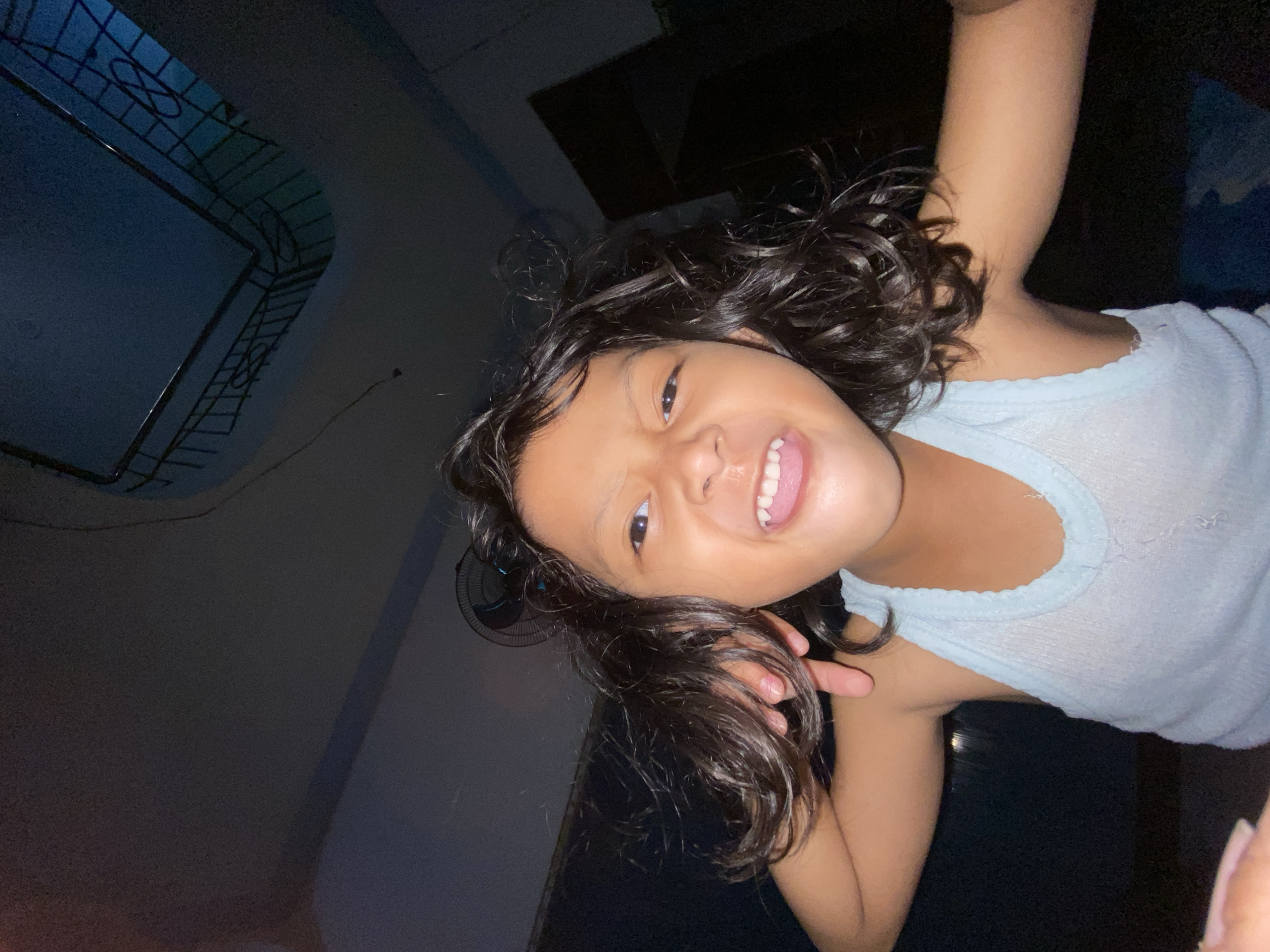 A young child with curly dark hair is smiling broadly and looking directly at the camera. The child is wearing a light blue tank top. The background is dark. A window with a metal grate is visible in the upper left corner, indicating an indoor location. The environment appears to be a home interior in Gelam, Indonesia, based on the reference location provided.