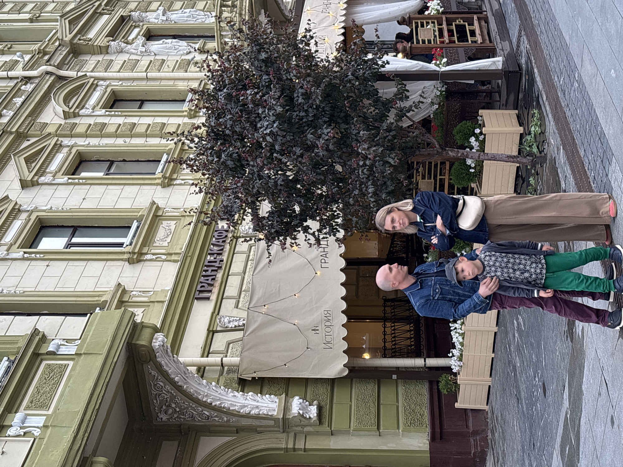 The image shows a family of three standing outside a building with ornate architectural details. A man, a woman, and a young boy are the main subjects. The man is wearing a denim jacket and is holding the boy. The woman is also wearing a denim jacket and is standing next to them. The boy is wearing a patterned shirt and green pants. They appear to be posing for a photo.
Behind them is a building with a light-colored facade adorned with intricate white moldings and sculptures. A large, leafy tree with dark foliage stands in front of the building. A cafe awning with string lights is visible, and there are some people seated at tables in the background, suggesting the setting is a public space, possibly a street or a plaza. The ground appears wet, hinting at recent rain or a damp atmosphere.
Visible text on the awning reads "ГРАНД КАФЕ" and "ИСТОРИЯ". The building's architecture and the Cyrillic text suggest a location in Russia. The lighting in the image is somewhat subdued, indicating it might be overcast or late afternoon. The overall mood appears casual and familial.