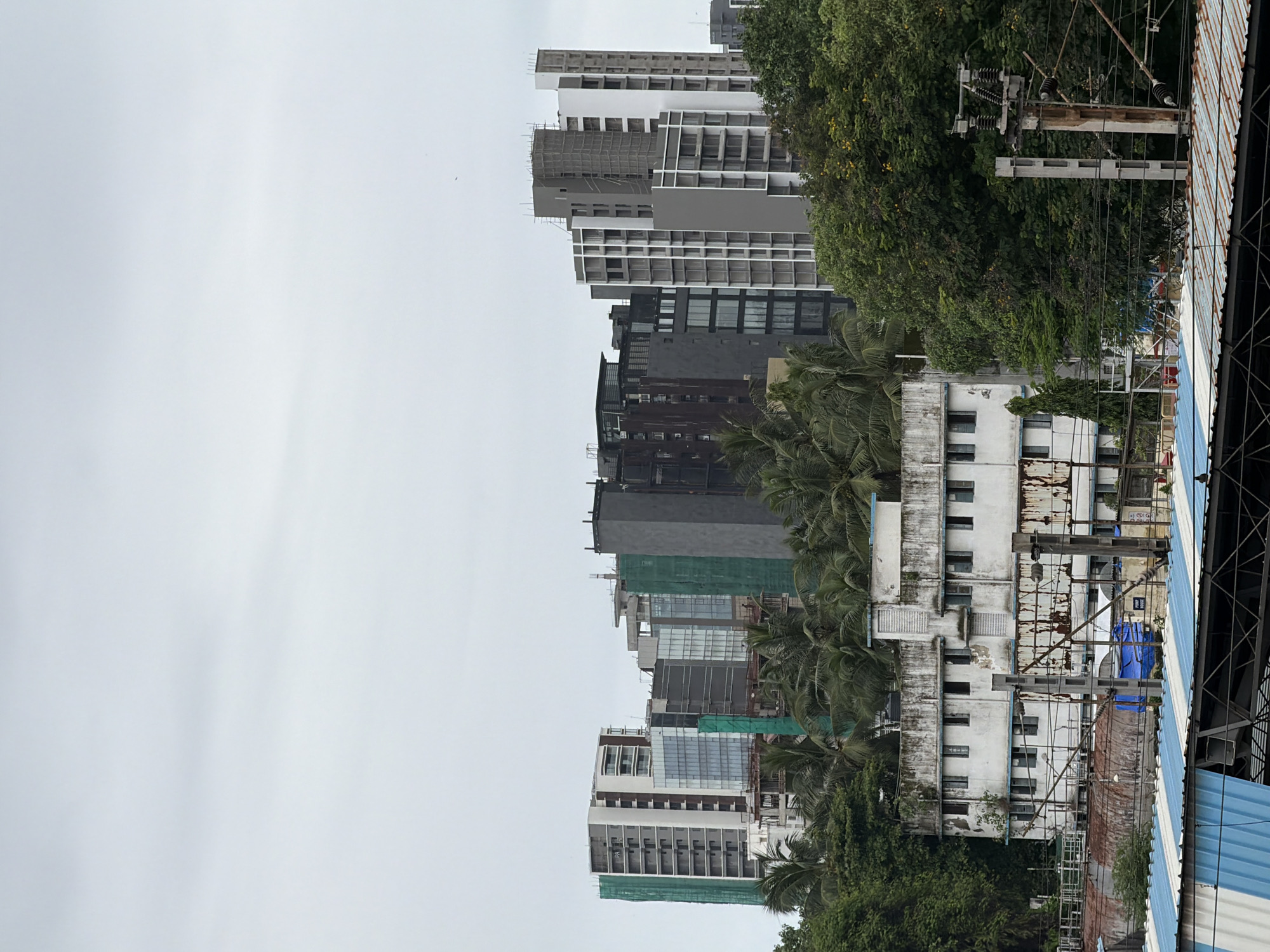 The image captures a view of a city skyline with multiple buildings of varying heights and architectural styles, set against an overcast sky. In the foreground, there is a lower, older white building with visible signs of wear and tear, featuring rows of dark windows and a blue-trimmed roof. Adjacent to this building are lush green trees and palm trees. Behind the older structure, a cluster of modern high-rise buildings, some appearing to be under construction with visible scaffolding and green netting, dominate the mid-ground and background. The architecture of these taller buildings includes elements of glass, concrete, and metal. In the far background, hints of more distant structures can be seen. The lower section of the image reveals the corrugated roof of a structure, possibly a market or transport hub, with blue and white stripes, and further below, a rusted metallic surface. Electrical poles and wires are also visible, suggesting an urban setting. The overall impression is one of urban development where older infrastructure coexists with new construction.