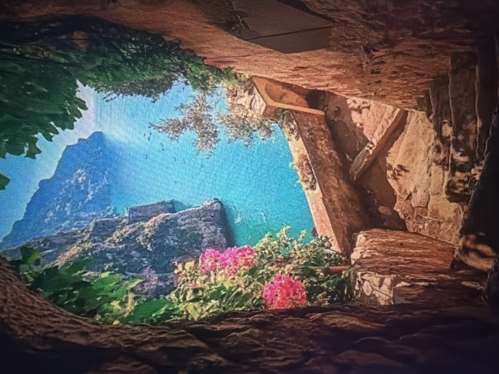 The image depicts a view through a rocky opening, possibly a cave or a narrow passage, looking out towards a vibrant turquoise sea and rugged cliffs. In the foreground, rough-hewn stone steps descend into shadow. Lush green foliage and bright pink flowers frame the opening on the left. To the right, the rocky walls of the passage are visible, textured and reddish-brown. The sea is dotted with several small boats. The scene suggests a scenic coastal location, potentially a path leading down to the water's edge. The city of Baan Mae Nam, Thailand, is not discernible in this image.
