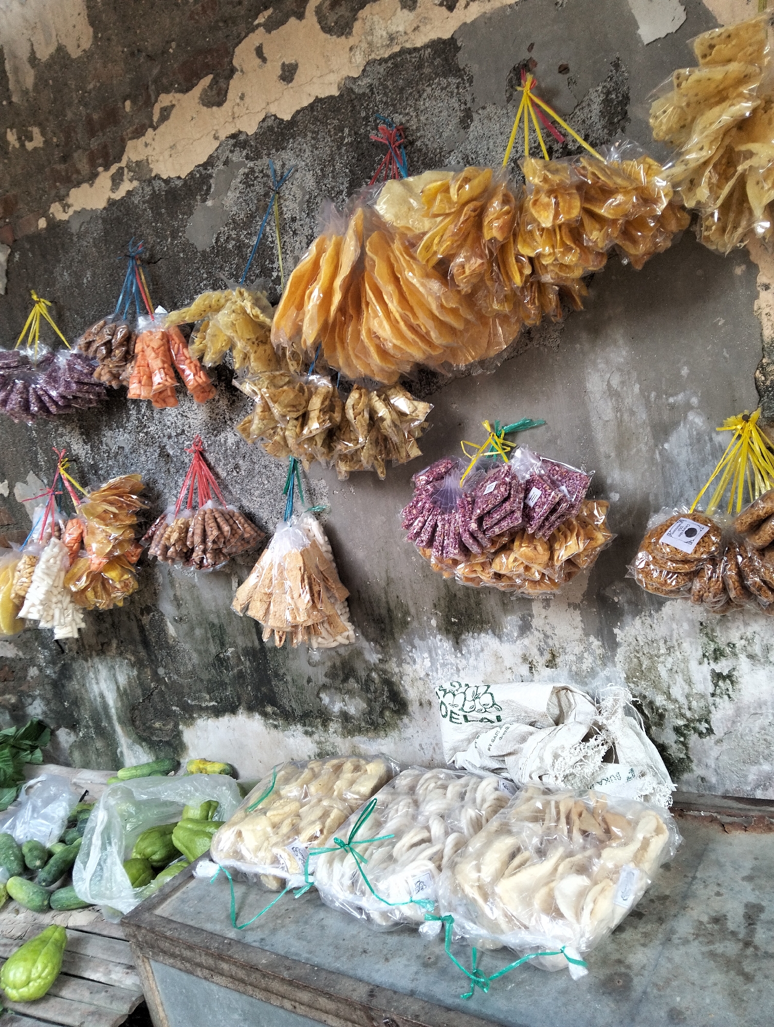 The image depicts a street stall in Muncar, Indonesia, showcasing a variety of dried snacks and fresh produce displayed against a weathered, textured wall. Hanging from strings attached to the wall are numerous plastic bags filled with assorted snacks. These include thinly sliced, pale yellow chips resembling krupuk, darker, irregularly shaped snacks that appear to be fried, and small, light brown crunchy items. Some bags contain purple-hued snacks, possibly flavored with spices or a coloring agent.
In the foreground, a low table or stall surface holds more packaged snacks, some of which are a creamy white color and others a light yellow. To the left of the table, a collection of fresh produce is visible. These include several elongated green cucumbers and what appear to be chayote fruits, some in a plastic bag and one prominently placed in the lower left corner. The overall scene suggests a local market or roadside vendor offering a mix of traditional Indonesian snacks and fresh vegetables. The wall behind the stall exhibits signs of age, with peeling paint and some green moss or algae growth, characteristic of an outdoor environment.