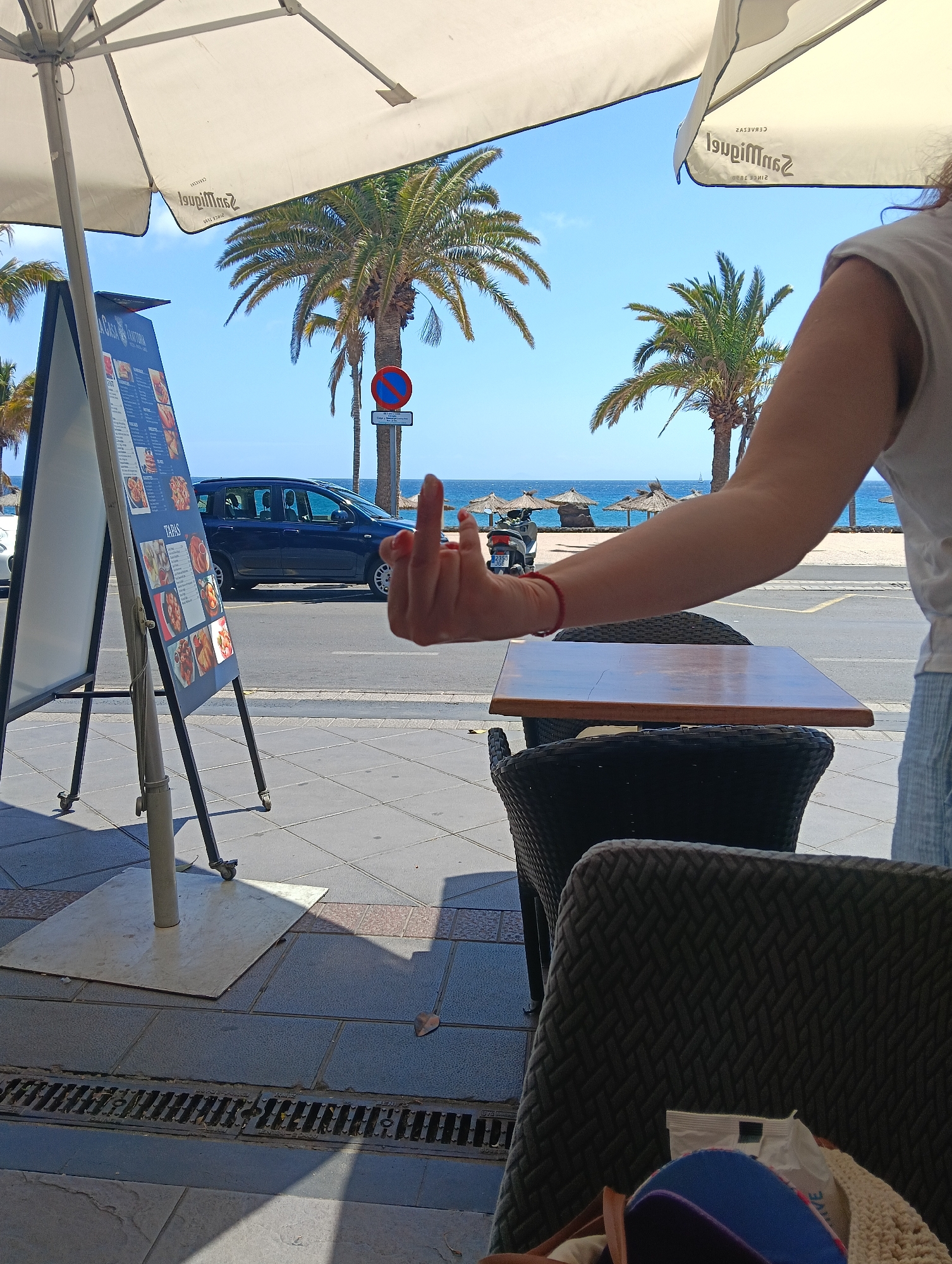 An outdoor, eye-level photograph captures a person's arm extending a middle finger towards the left, set against a vibrant coastal backdrop in Puerto Del Carmen, Spain. The person, partially visible on the right with a white t-shirt and blue patterned shorts, wears a red bracelet on their wrist. In the foreground, part of a wooden table and two dark wicker chairs indicate an outdoor cafe setting. A blue cap and a white bag are visible in the nearest chair.
To the left, a white menu board on wheels advertises "TAPAS" with various food pictures. Overhead, a large white parasol, partially open, shows the mirrored text "San Miguel" and "CERVEZAS SINCE 1890." Beyond the cafe, a paved street features a dark blue car and a scooter (license plate "2385 JGE") parked by the curb. Further in the background, a pristine blue sky meets the ocean, where several straw beach umbrellas line the sandy shore. Lush palm trees are scattered throughout the scene, and a red circular "no parking" sign is visible near one of the trees. The bright sunshine suggests a clear, warm day, likely mid-day, casting shadows from the umbrella and objects. The overall scene depicts a casual, sunny day at a Spanish coastal resort, punctuated by the defiant or playful gesture of the extended middle finger.