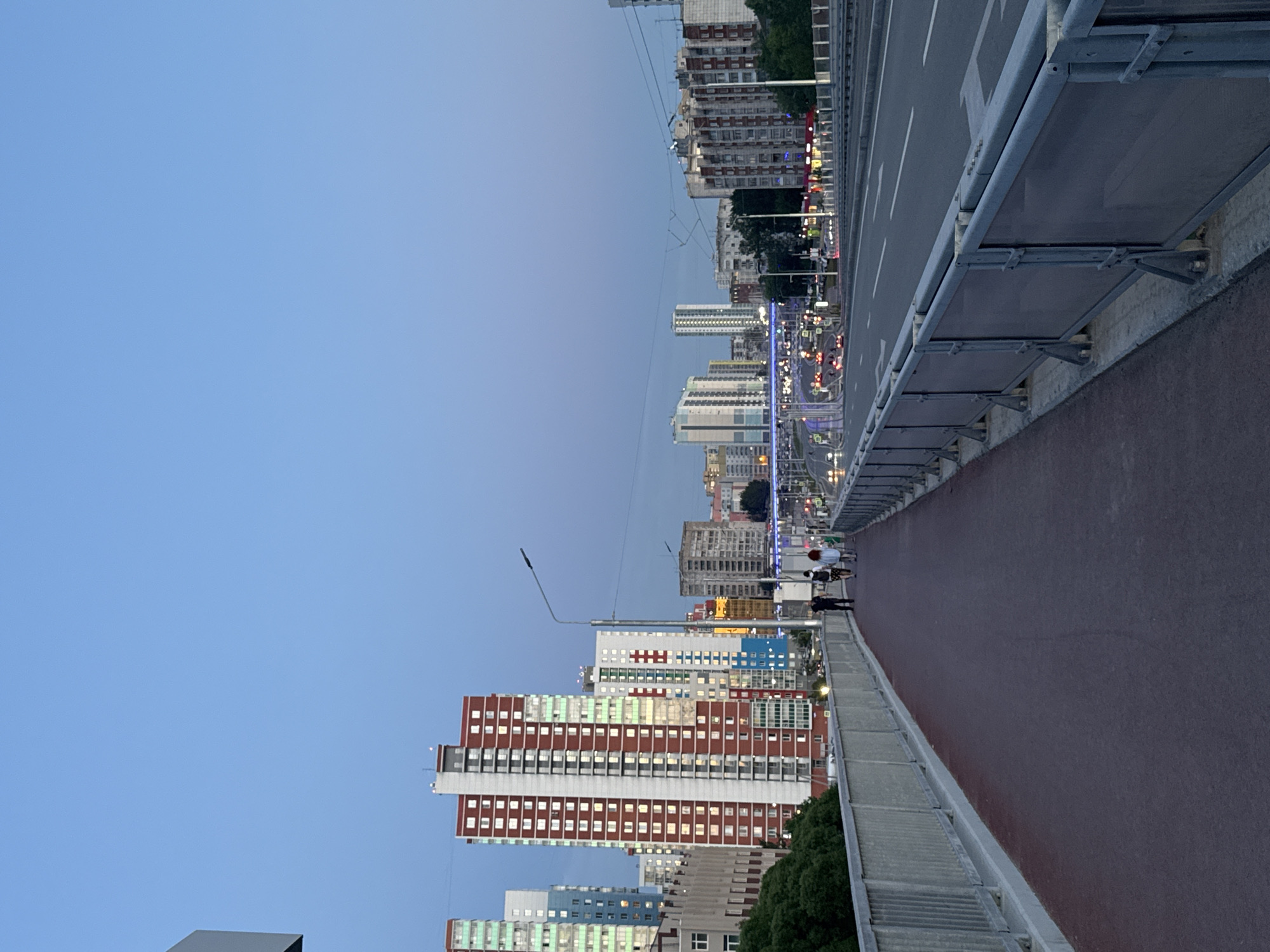 The image shows an elevated perspective of a cityscape during twilight. A pedestrian walkway, with a reddish-brown surface, runs alongside a roadway. Buildings of varying heights and colors line the street below. The sky is a gradient of blue. Several people are walking on the walkway. The location is likely Perm, Russia based on the architecture and layout of the city.