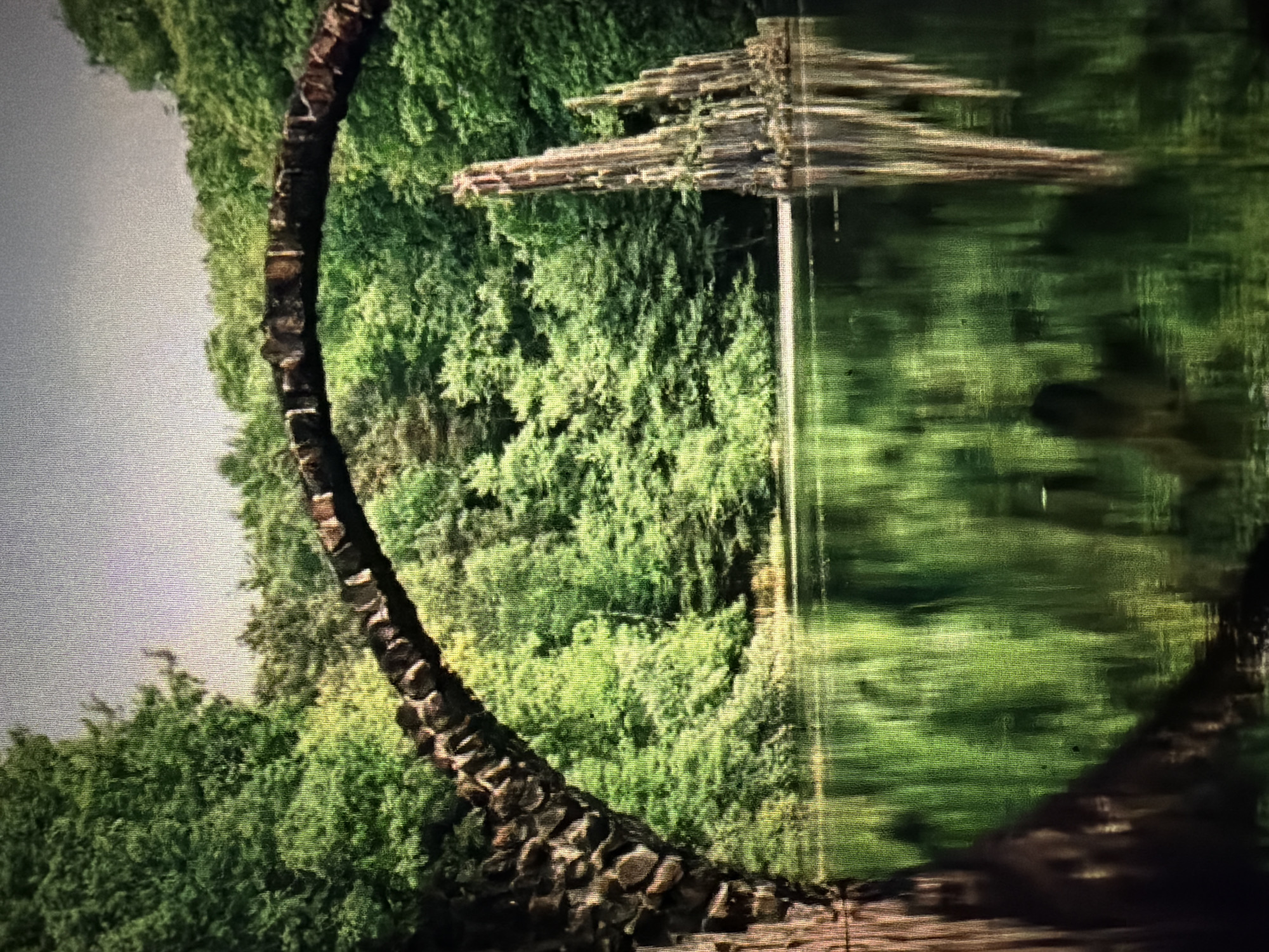 The image depicts the Rakotzbrücke, a historic arched bridge made of stone, set within a lush forest. The bridge gracefully spans a calm body of water, creating a near-perfect reflection of its structure and the surrounding greenery. To the right of the bridge, a distinctive rock formation rises from the water, resembling a fantastical spire or a collection of weathered columns. The water's surface is smooth, mirroring the dense foliage of trees that form a backdrop, with the sky visible as a pale, muted expanse above. The scene evokes a sense of tranquility and natural beauty, with the man-made bridge blending harmoniously with the wild, verdant landscape. This specific location is in Kromlau Park, Germany, not Baan Mae Nam, Thailand.