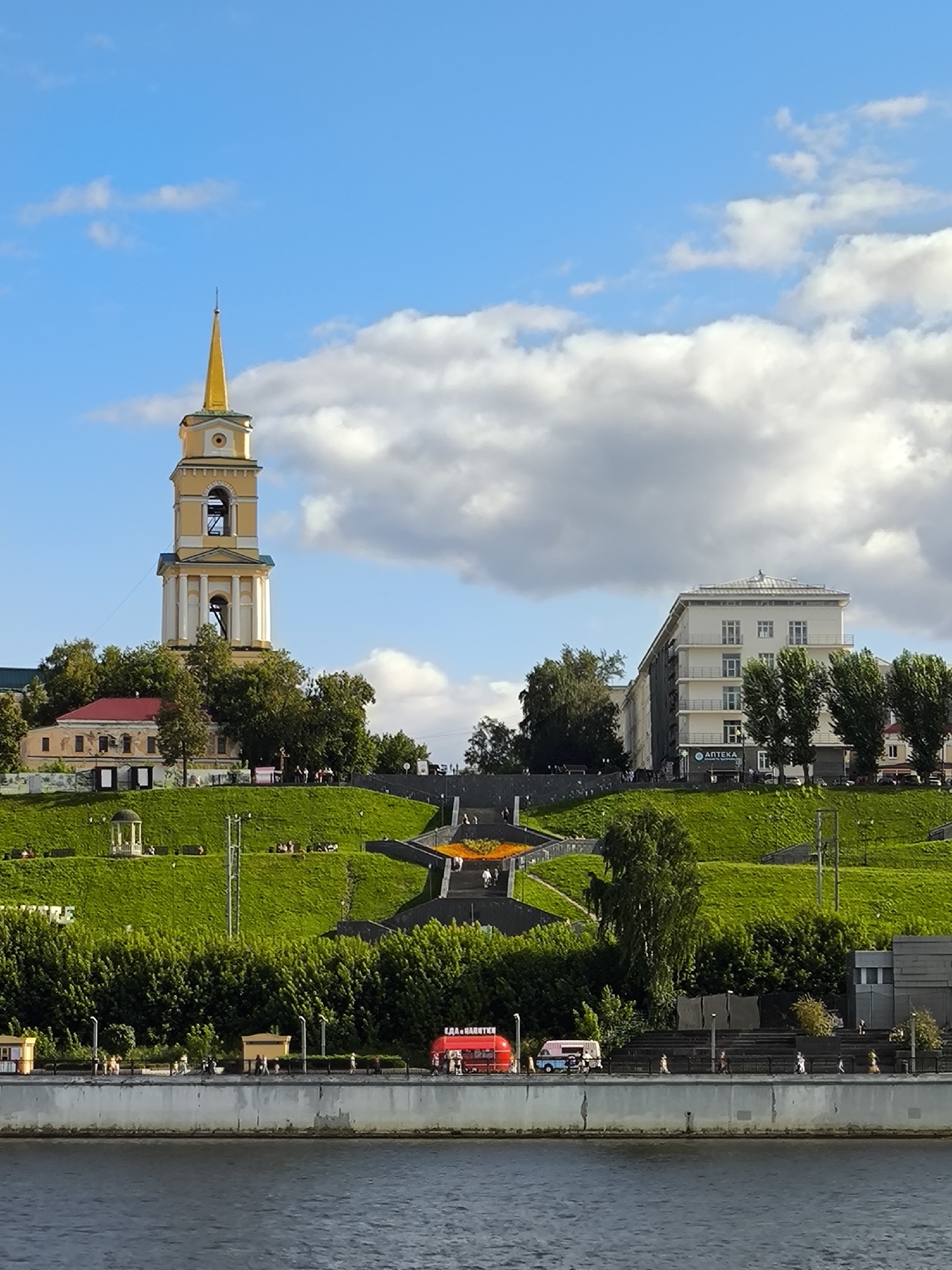 This image captures a view of the Perm embankment in Perm, Russia, on a sunny day with some clouds. A prominent yellow bell tower with a golden spire stands on a grassy hill. Adjacent to the bell tower is a light-colored, multi-story building. A set of wide stairs with illuminated accents leads up the grassy slope, with people visible walking on it. The foreground features a body of water, likely the Kama River, with a concrete embankment along its edge. Along the embankment, a red food truck and a light blue and white food truck are parked, with several people walking nearby. Dense green trees line the base of the hill, separating it from the embankment.