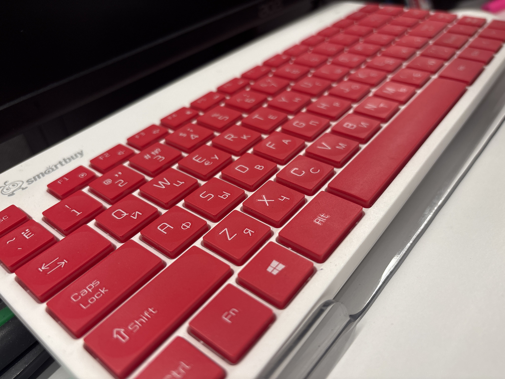 A close-up, high-angle shot shows a red and white keyboard with a distinctive "smartbuy" logo on the left side. The keyboard is angled, with the top right of the image blurred. The keys are red with white lettering, and the base of the keyboard is white. Some of the keys are visible with individual characters and symbols, such as "F1", "@ 2", "W", "E", "R", "T", "Y", "U", "I", "O", "P", "A", "S", "D", "F", "G", "H", "J", "K", "L", "Z", "X", "C", "V", "B", "N", "M", "Caps Lock", "Shift", "Fn", and the Windows logo. The keyboard appears to be on a desk or table, with a dark, out-of-focus background that could be a laptop screen. The lighting is bright, creating some glare on the keys. No people are visible in the image. The context of Birobidzhan, Russia, is not visually represented in the image itself.