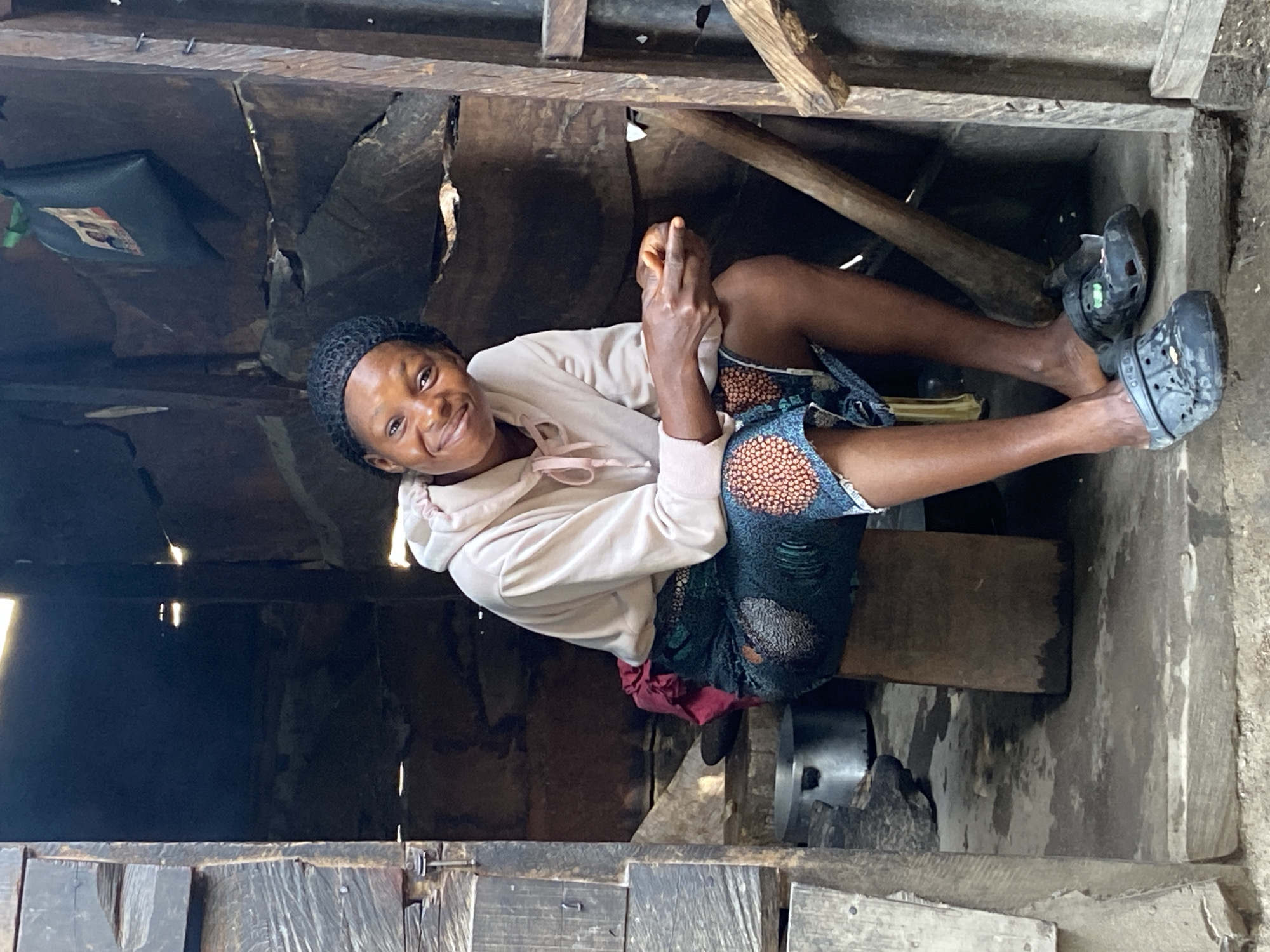 A smiling woman sits in a rustic dwelling. She wears a light pink hooded sweatshirt, a dark patterned skirt, and black Crocs. The structure appears to be made of wood and other rudimentary materials. A black container hangs from the ceiling, while a wooden beam and other objects are scattered around the room. The context provided indicates this scene takes place in Mararaba, Nigeria.