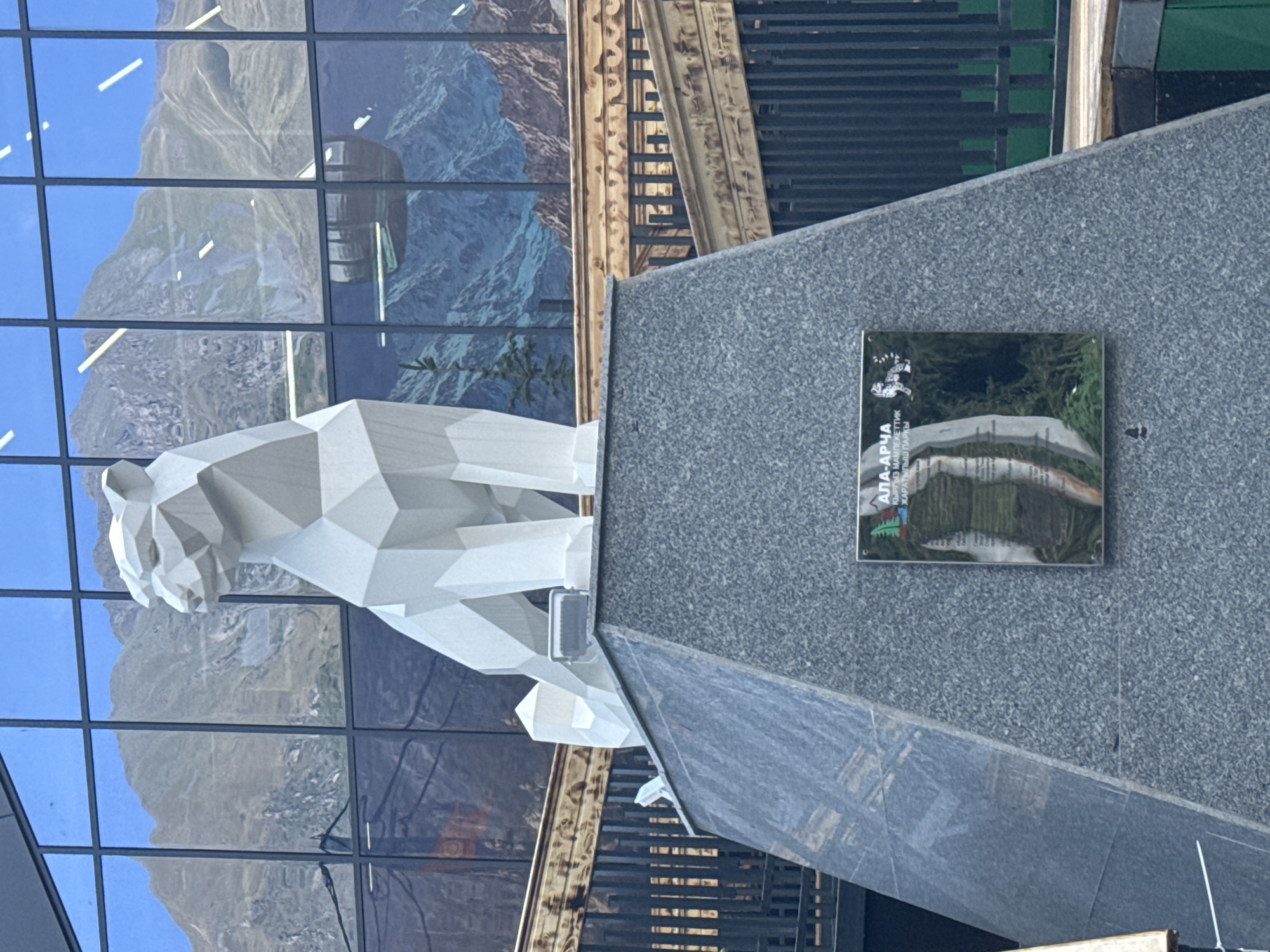 A white, geometric sculpture of a snow leopard is prominently displayed on a granite pedestal in front of a large window. The window reflects a mountain landscape with a cable car, suggesting an outdoor or mountainous location, possibly a tourist attraction. A plaque is affixed to the pedestal, bearing text and an image of a snow leopard. The background includes wooden railings and metal fences. The overall impression is one of a public monument in a scenic or recreational area.