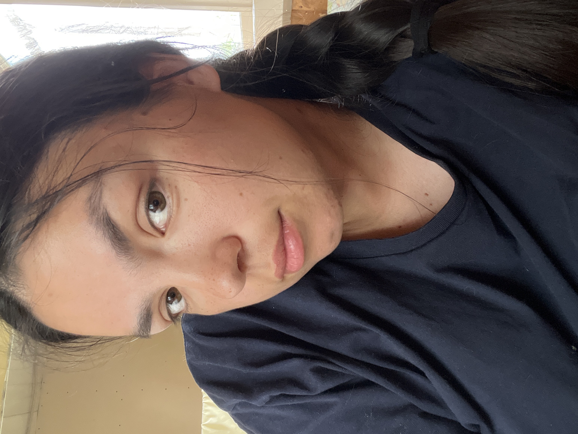 A young woman with dark hair, styled in a braid resting over her left shoulder, is depicted from the chest up. She has fair to medium skin with noticeable freckles on her nose and cheeks. Her dark brown eyes are focused directly on the viewer. Her full lips are slightly parted, revealing a light pink tone. She is wearing a plain, dark navy blue crew-neck t-shirt. The setting is indoors, with a light-colored wall or surface visible on the left side of the frame. To the right, a bright window with a light-colored frame indicates a daytime environment. The image contains no visual information to determine its geographical location as the city of Oktyabrsky, Russia, or any other specific city or country.