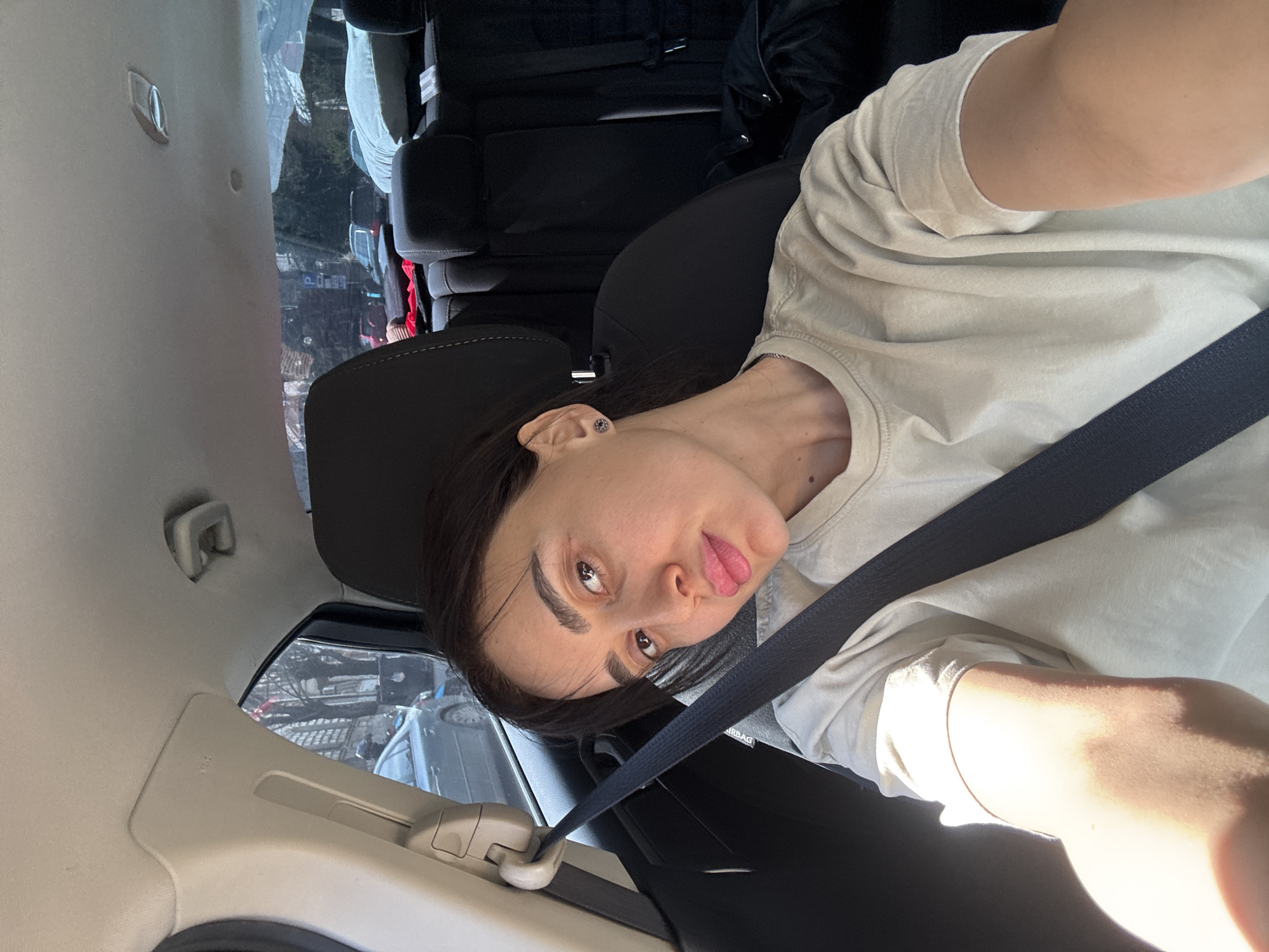 A woman is taking a selfie while sitting in the passenger seat of a car. She is wearing a light-colored t-shirt and a dark seatbelt. The interior of the car is visible, including the seats and window. Outside the window, a street scene suggests a location in Tbilisi, Georgia. The street is lined with buildings and cars.