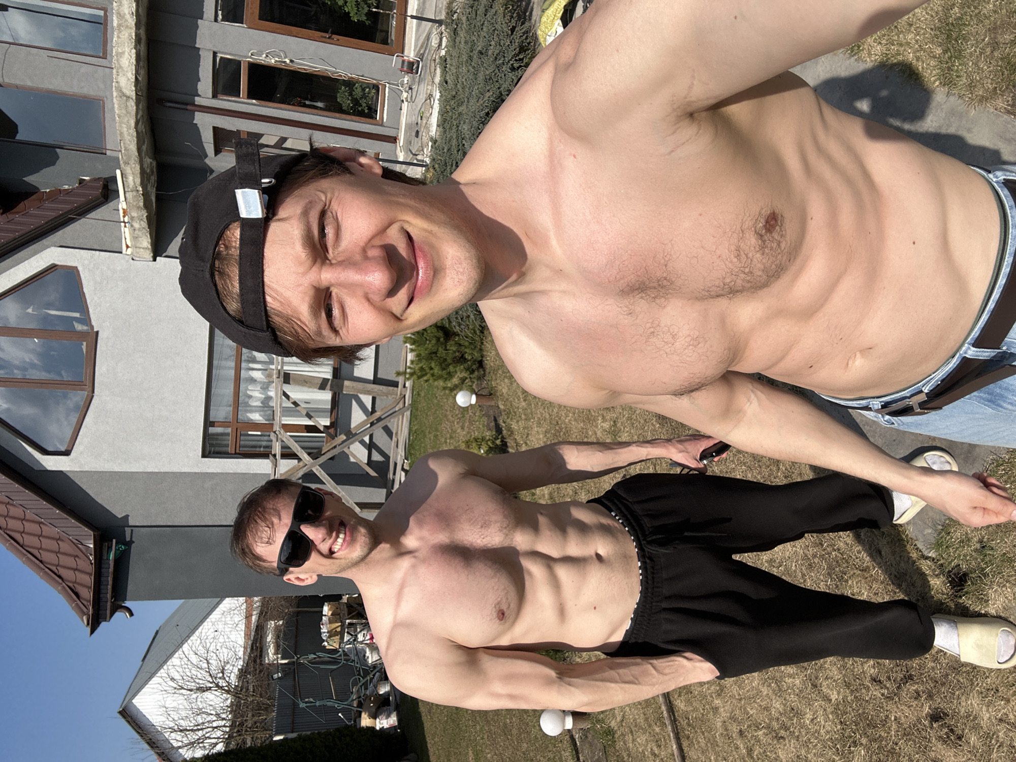 Two shirtless men are posing for a selfie outdoors on a sunny day. The man in the foreground wears a black baseball cap and smiles. The man behind him wears sunglasses and black sweatpants with a white and black striped waistband, and is barefoot. They are standing in a yard with grass, near a house. The specific location in Kursk, Russia, cannot be determined from the image.