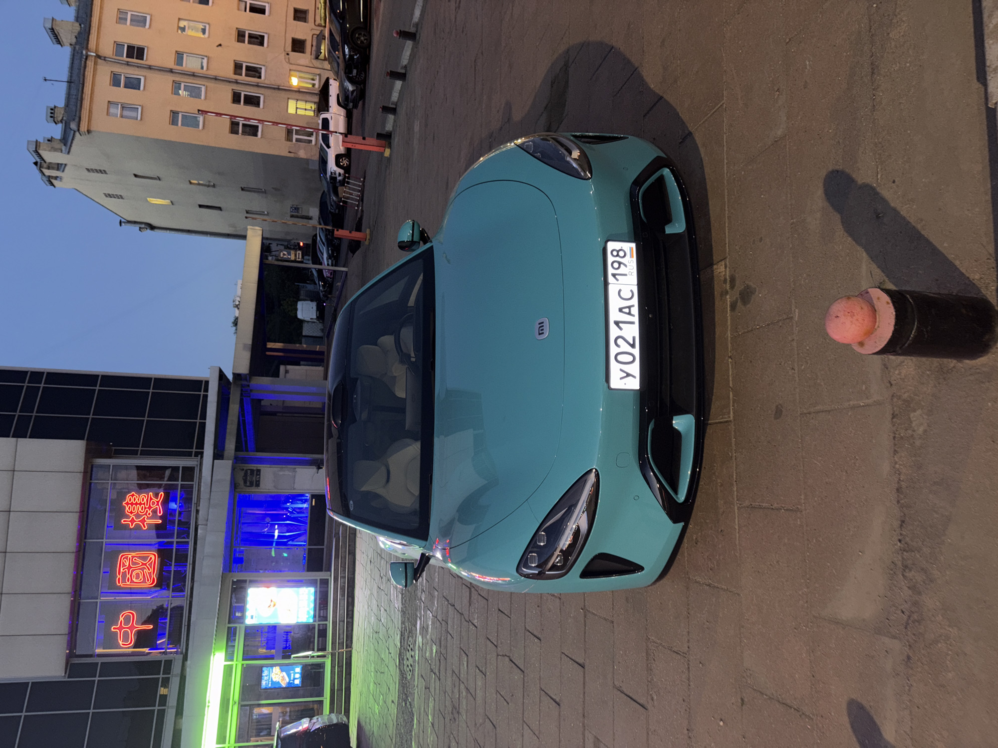 A teal-colored electric sedan with a black roof is parked on a cobblestone street in Saint Petersburg, Russia. The car is facing the camera, and its license plate reads "Y021AC 198". In the background, there is a modern building with Chinese characters illuminated by neon lights, suggesting a restaurant or establishment. Adjacent to it, a multi-story building with several windows is visible. The sky is dark, indicating it is either dusk or night. The scene appears to be a public street in an urban environment.