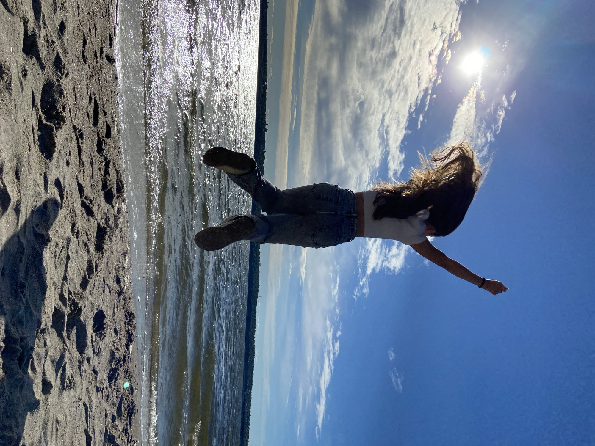 A young person with long, dark hair is captured mid-air, jumping on a sandy beach. They are wearing a white top and blue jeans. Their arms are raised, with their right arm extended upwards. The sun is shining brightly in the blue sky, with some clouds present. The ocean is visible behind them, with waves gently lapping at the shore. The image conveys a sense of freedom and joy. The location is the city of Unknown, Unknown.