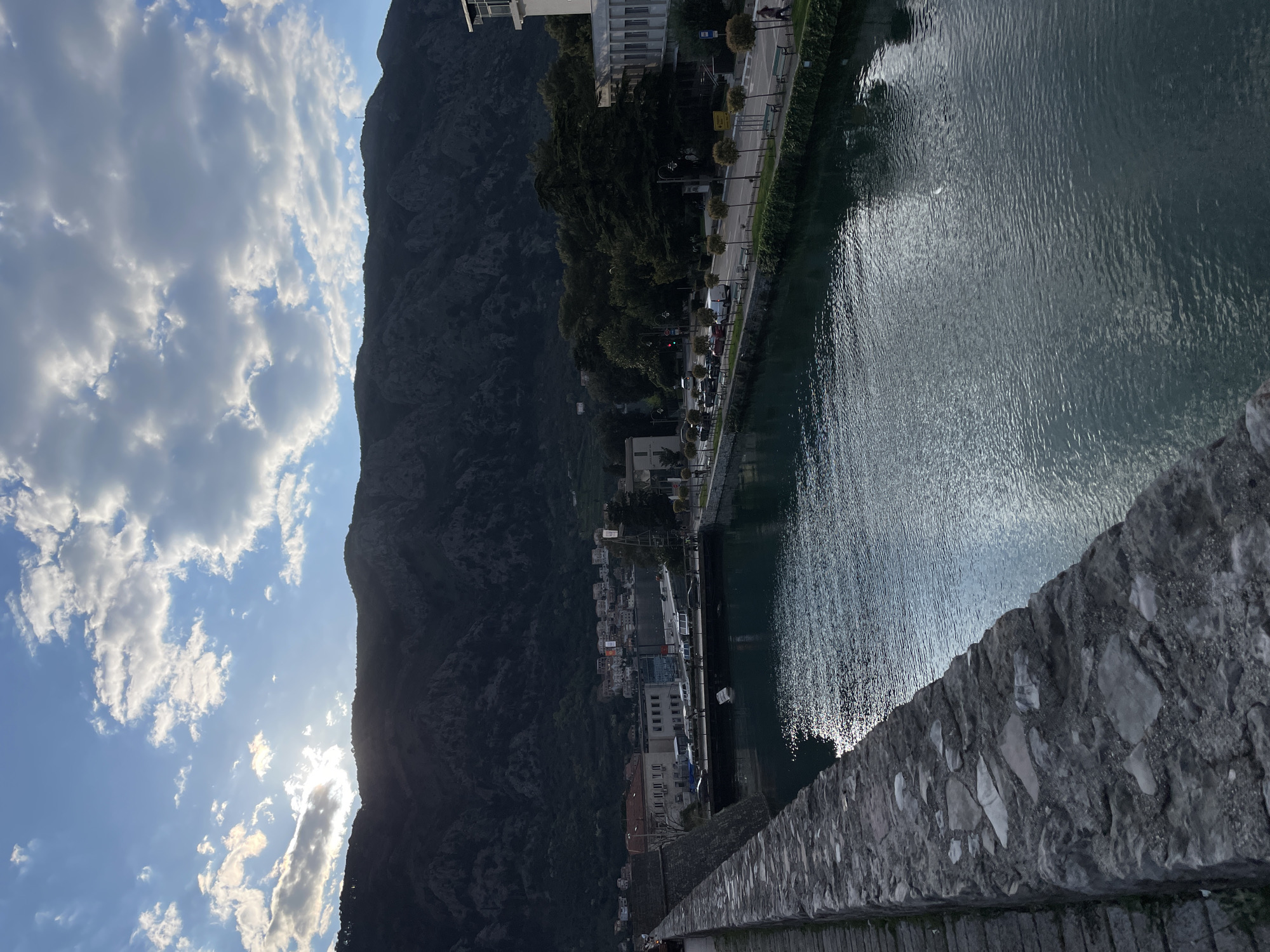 The image depicts a view from a stone bridge in Kotor, Montenegro. To the left, a large, dark mountain rises next to a blue sky filled with scattered white clouds. Below the mountain, there is a town with buildings of varying sizes and colors, nestled along the edge of the river. A paved road runs parallel to the river with trees and vehicles visible. The river fills the right side of the frame with sunlight reflecting off the water's surface. The foreground is dominated by a grey stone wall, likely part of the bridge's structure.