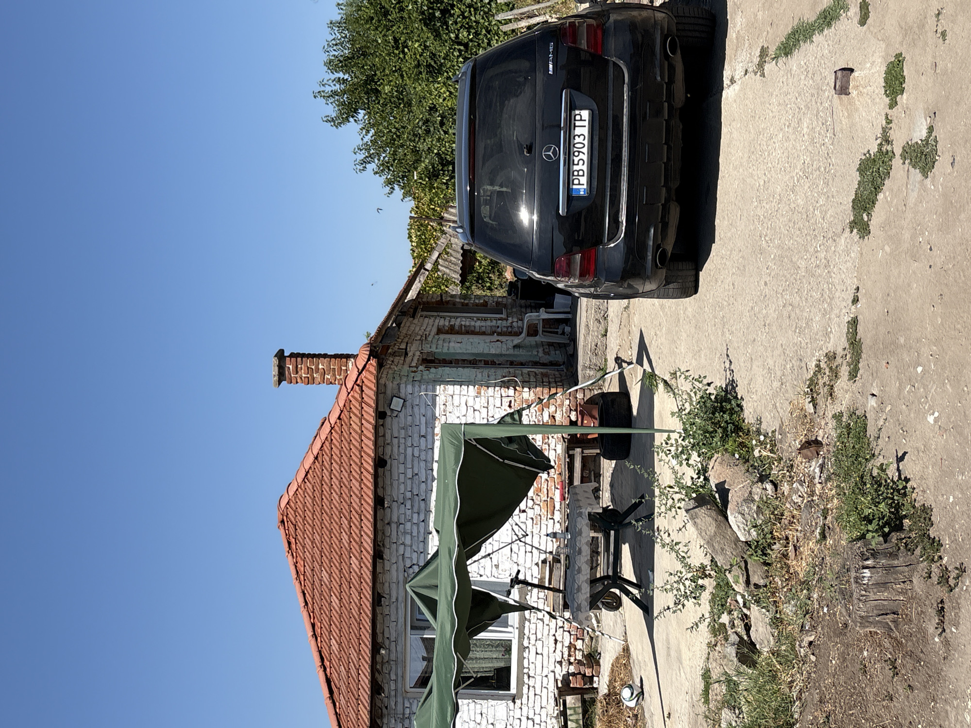 The image depicts a sunny outdoor scene at a residential property in Voysil, Bulgaria. In the foreground, a black Mercedes-Benz SUV with license plate PB5903TP is parked on a concrete surface. To the left of the car, a white brick house with a red tiled roof is visible. In front of the house, a green canopy tent is set up over a table with chairs, and a soccer ball rests on the ground. The environment appears somewhat unkempt, with overgrown vegetation and debris scattered around the yard. The sky is clear and blue.
