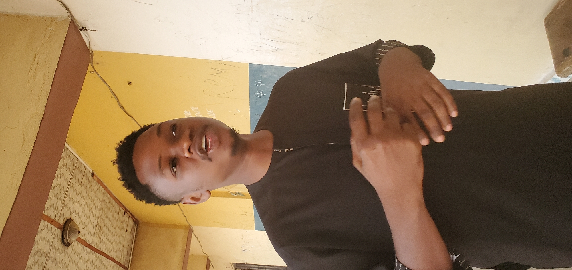 A man stands indoors in Mararaba, Nigeria. He is dark-skinned with short, dark hair and a beard. He wears a black shirt with a white rectangular detail on the chest. His hands are raised. The background shows walls painted yellow and white with some writing on one section. A portion of a window is visible at the bottom. The lighting in the room is subdued.