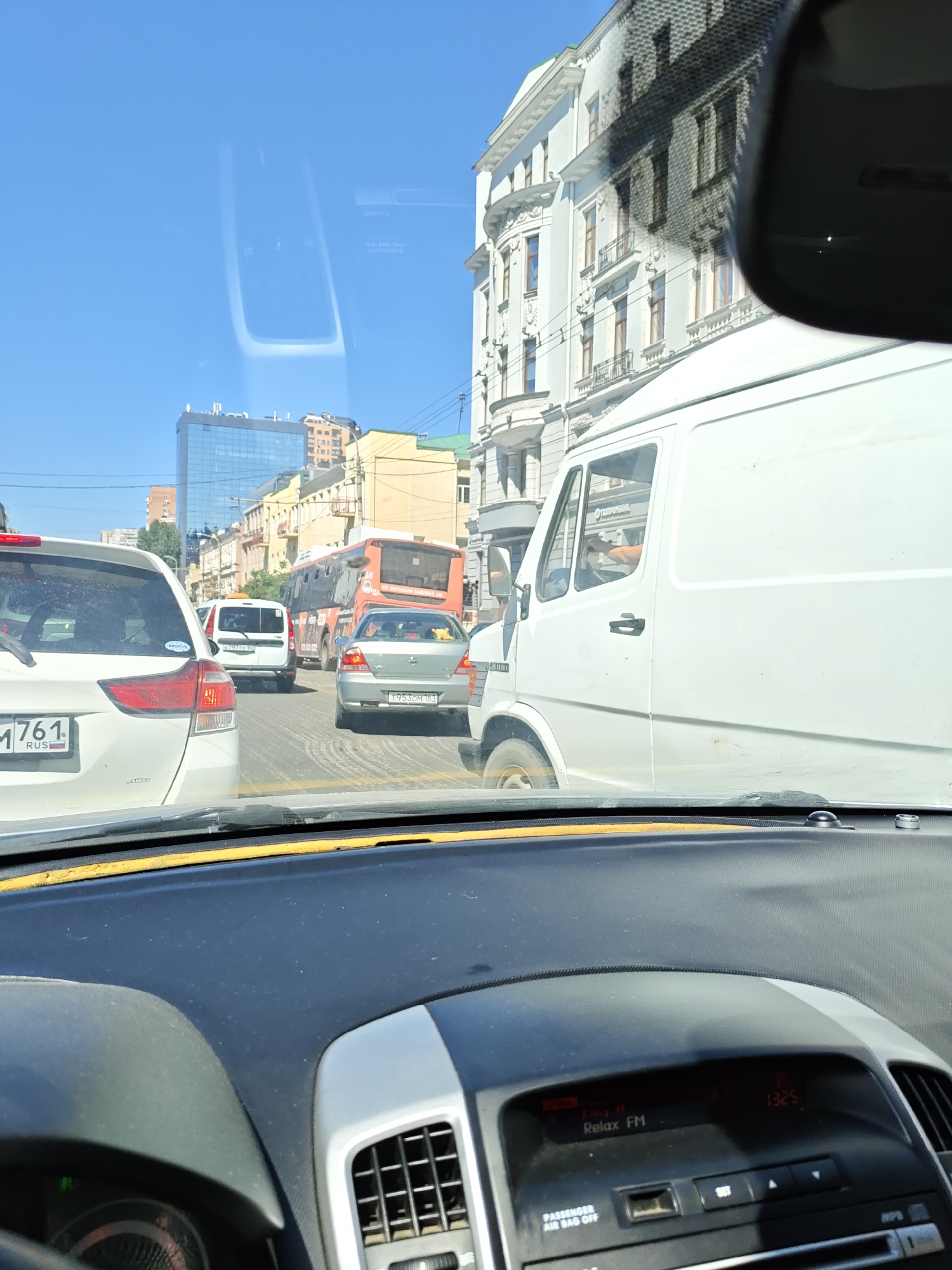 The image is taken from inside a car, looking through the windshield at a traffic jam on a sunny day. Multiple cars are visible, including a white SUV in the foreground, a white van, a silver sedan, and an orange bus. The buildings in the background are a mix of old European-style architecture and modern glass skyscrapers. A reflection of the sky and some of the car's interior, including part of the dashboard and rearview mirror, are visible on the windshield. The dashboard shows a radio display with "Relax FM" and the time "13:25". License plates are visible on some vehicles, with one in the foreground reading "M 761 RUS". The overall scene depicts a typical urban traffic situation.