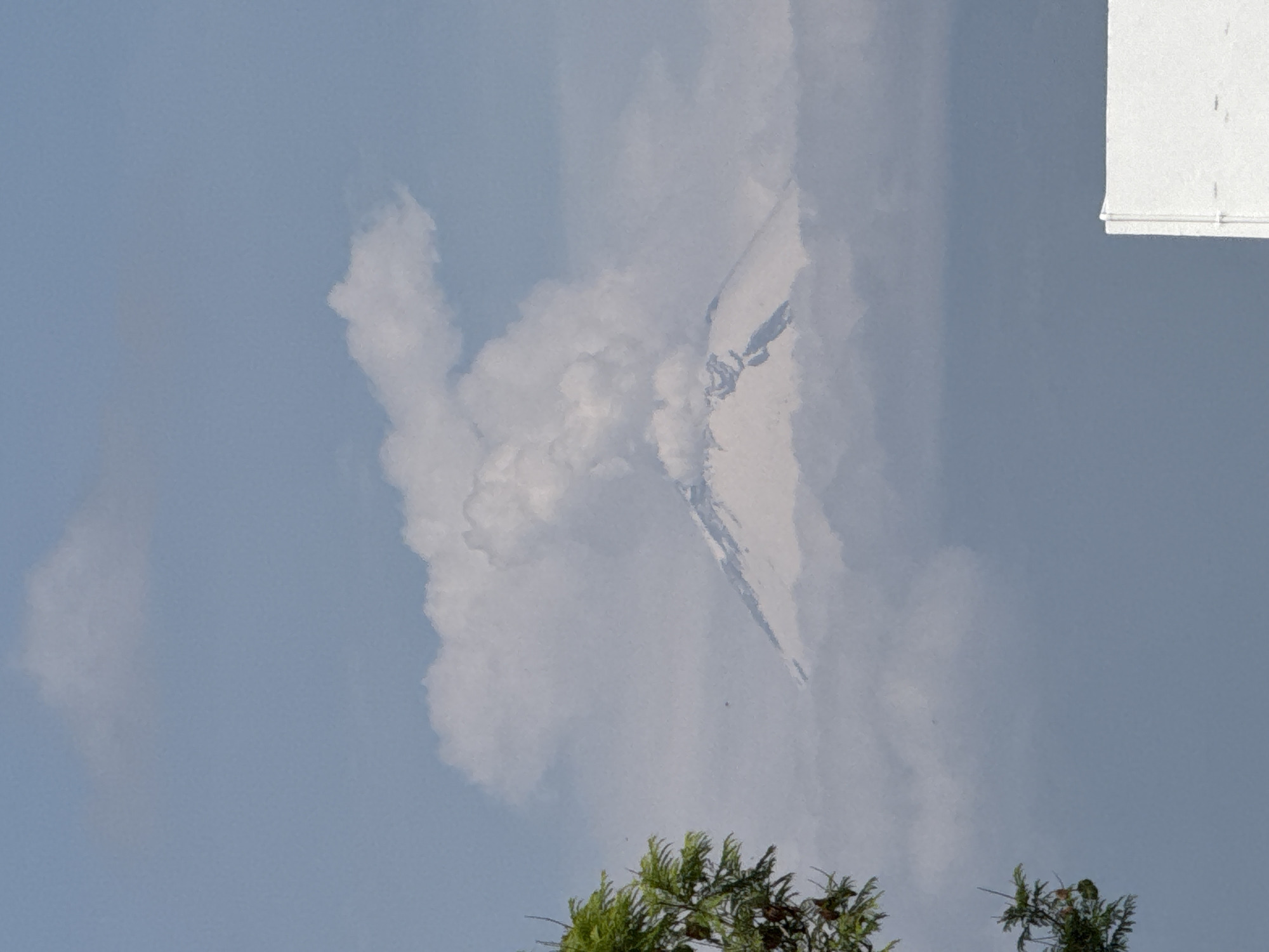 The image shows a daytime sky over San Bernardino Tlaxcalancingo, Mexico. Dominating the view are clouds, some resembling a mountain peak, and a blue sky. Green trees are visible in the foreground. A white structure is in the upper right corner.