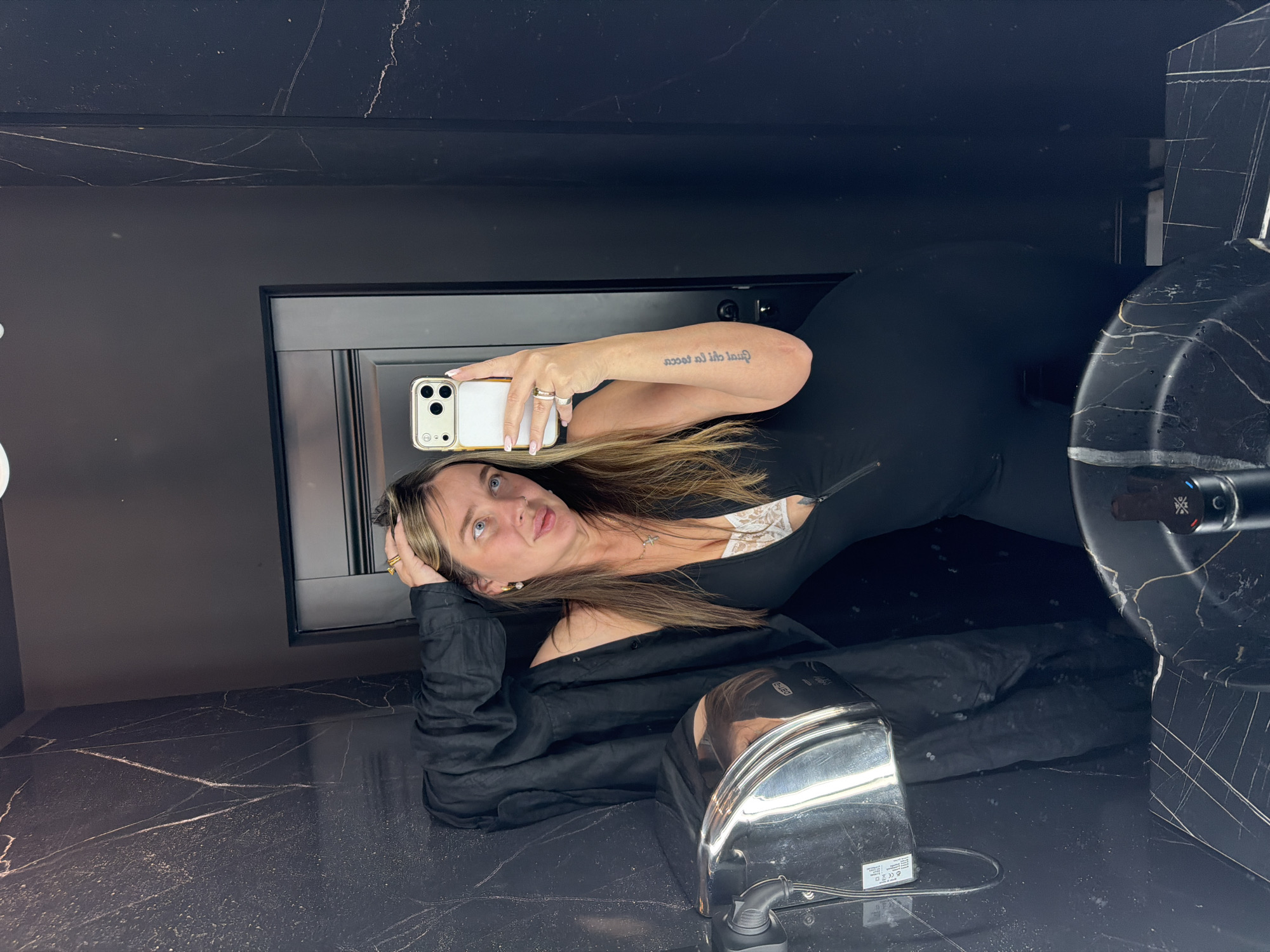 A woman is taking a selfie in a bathroom with black marble countertops and walls with white veining. She is wearing a black top and has long, light brown hair. Her phone is white with a gold bumper. A hand dryer is visible on the countertop. The specific location in Moscow, Russia cannot be determined from the image.