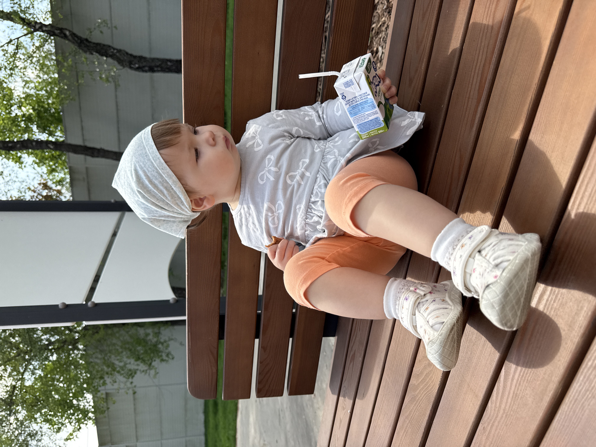 A child sits on a wooden bench, holding a juice box with a straw, with foliage and a gray, open-air structure in the background, suggesting a park or recreational area in Moscow, Russia.