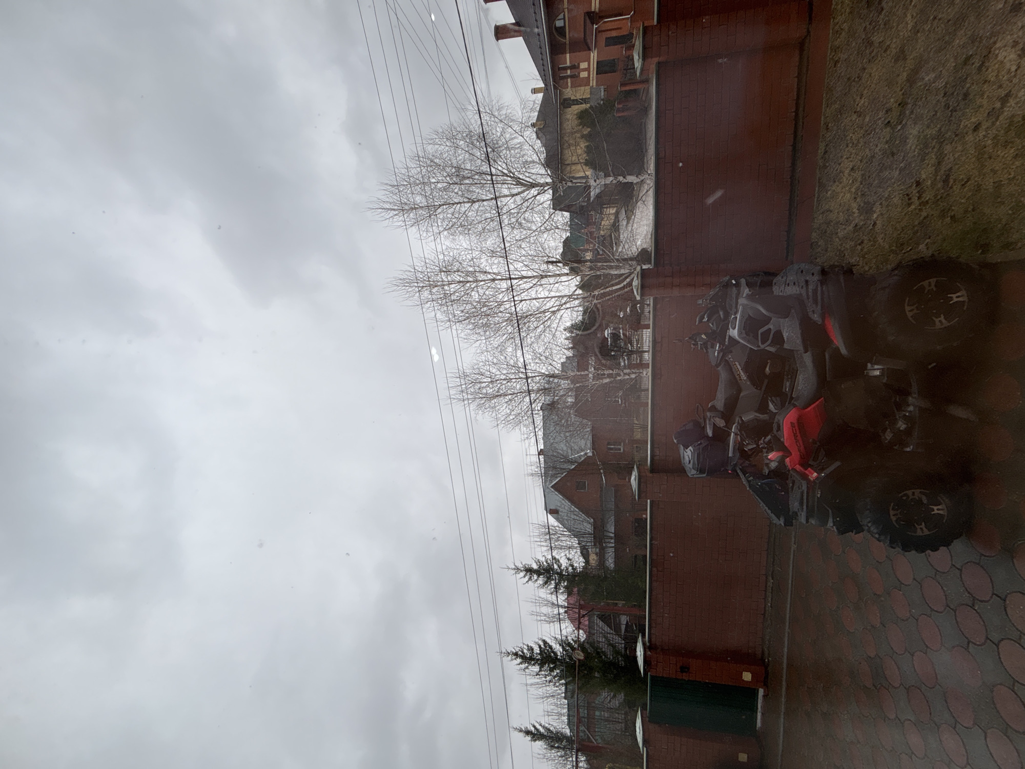 The image shows an outdoor scene in Ferma, Russia. The sky is overcast. A black and red all-terrain vehicle is parked on a red brick-paved area adjacent to a red brick wall. Beyond the wall is a residential area with houses, trees, and power lines. It appears to be a snowy or rainy day.