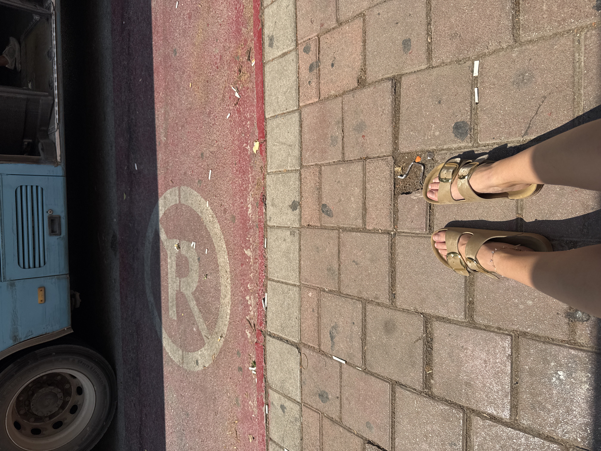 The image depicts a street scene, likely a bus stop, with a blue bus on the left. A red-painted asphalt surface with a white circular stencil marking is visible in the foreground, suggesting a designated area. The ground is paved with interlocking bricks, which are interspersed with cigarette butts, indicating litter. Two sand-colored sandals are worn by a person standing on the brick-paved sidewalk. The scene appears to be outdoors, under daylight. Based on the typical street infrastructure and the presence of a bus stop, this location could be in Antalya, Turkey.