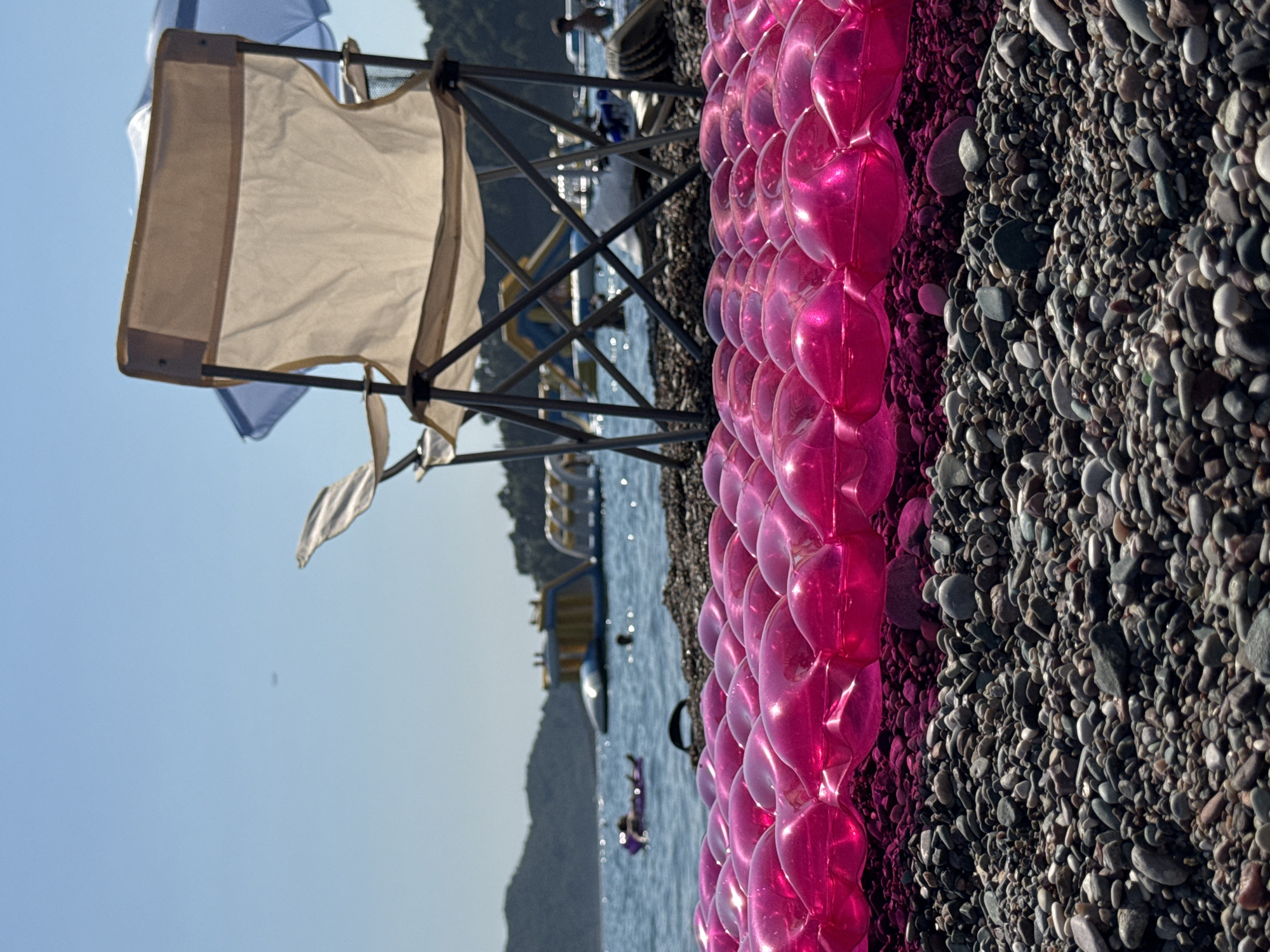 The image captures a sun-drenched scene on a pebble beach in Boreti, Montenegro. In the foreground, a bright pink inflatable mattress lies partially unfolded, its air-filled pockets reflecting the sunlight. The beach itself is covered in small, varied-colored pebbles.
Behind the mattress, a white canvas folding chair with a light brown trim stands prominently. To the left and right of the chair, parts of other chairs and a blue umbrella are visible, suggesting a comfortable seating area.
The background transitions to the calm, blue waters of the Adriatic Sea. Several people are enjoying the water; one person is seen relaxing on a purple inflatable in the middle distance, while another is partially visible near the water's edge. Further out, large inflatable water park structures with slides and platforms are floating in the sea, indicating recreational activities available in the area.
The horizon is marked by a lush, green, tree-covered hillside, characteristic of the mountainous coastline of Montenegro. The sky above is a clear, bright blue, with no clouds visible, reinforcing the impression of a warm, sunny day. The overall scene conveys a relaxed atmosphere associated with a beach vacation.