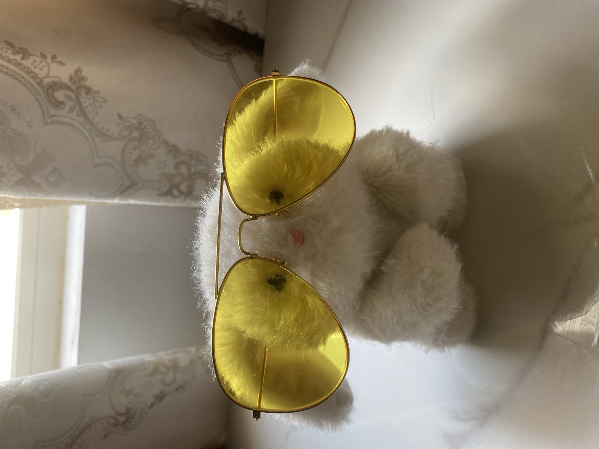 A fluffy white stuffed animal, resembling a rabbit, is positioned on a white, marble-like surface. It is wearing oversized, aviator-style sunglasses with bright yellow lenses. The sunglasses are a prominent feature, obscuring the stuffed animal's eyes. The background consists of sheer, patterned curtains and a window, suggesting an indoor setting. The lighting indicates natural light, possibly from the window. The scene is focused on the stuffed animal and its accessories. There is no indication of a specific location within the city of Thuận An, Vietnam.