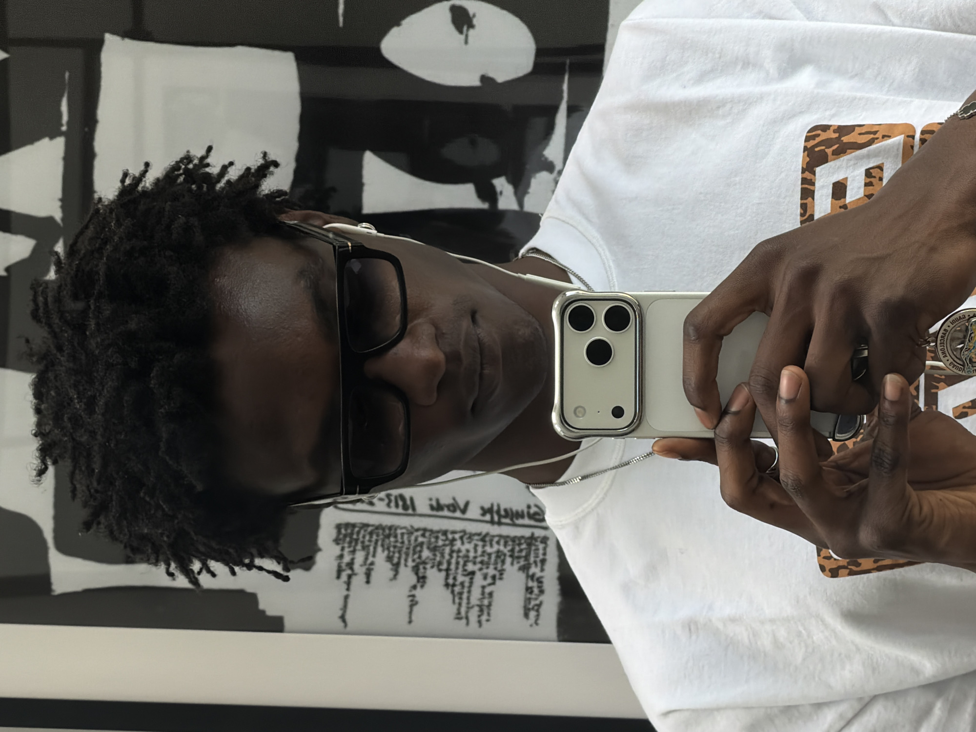 A dark-skinned young man is taking a mirror selfie. He has dark, curly hair, is wearing black sunglasses and white earbuds. He is holding a silver smartphone in both hands and wearing a white t-shirt with a graphic design on the front. He is also wearing a silver chain necklace. The background appears to be a wall with black and white abstract art. The image does not provide any specific details that would indicate the location is in London, United Kingdom.