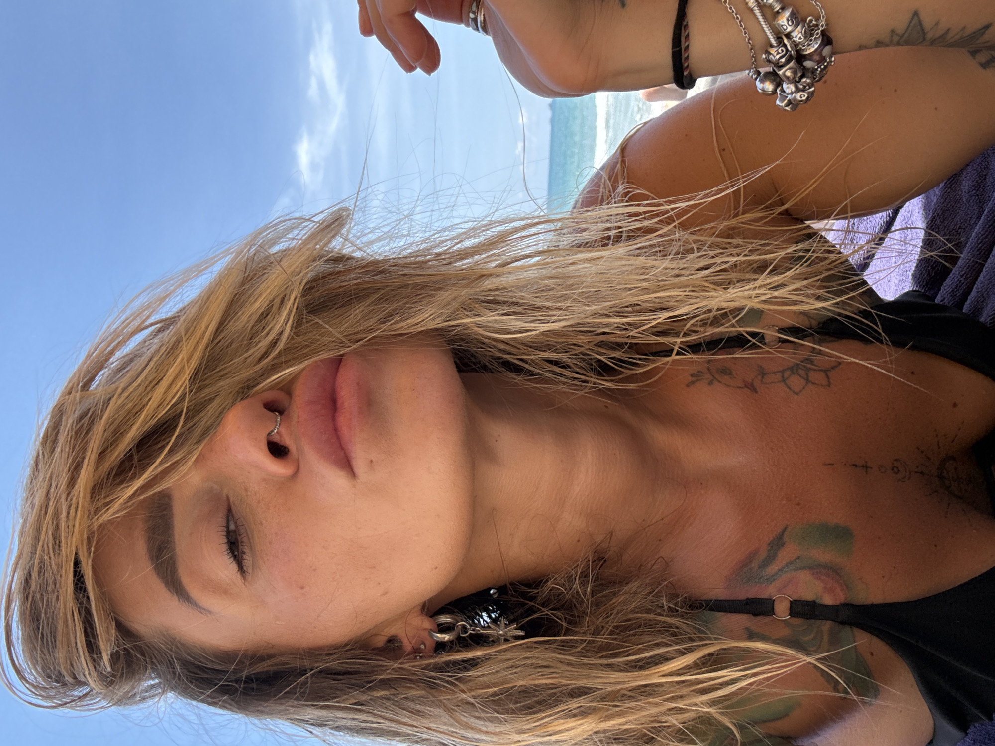 A close-up shot captures a woman with blonde, wet hair against a backdrop of the ocean and a clear blue sky, suggesting a beach location. She is wearing a black bikini top and has several tattoos visible on her chest and arm. She has a nose ring and is wearing earrings and bracelets. Her expression suggests she is posing for the camera, with her lips puckered. The overall impression is of a casual, beachside portrait. The location is identified as Choeng Thale, Thailand.