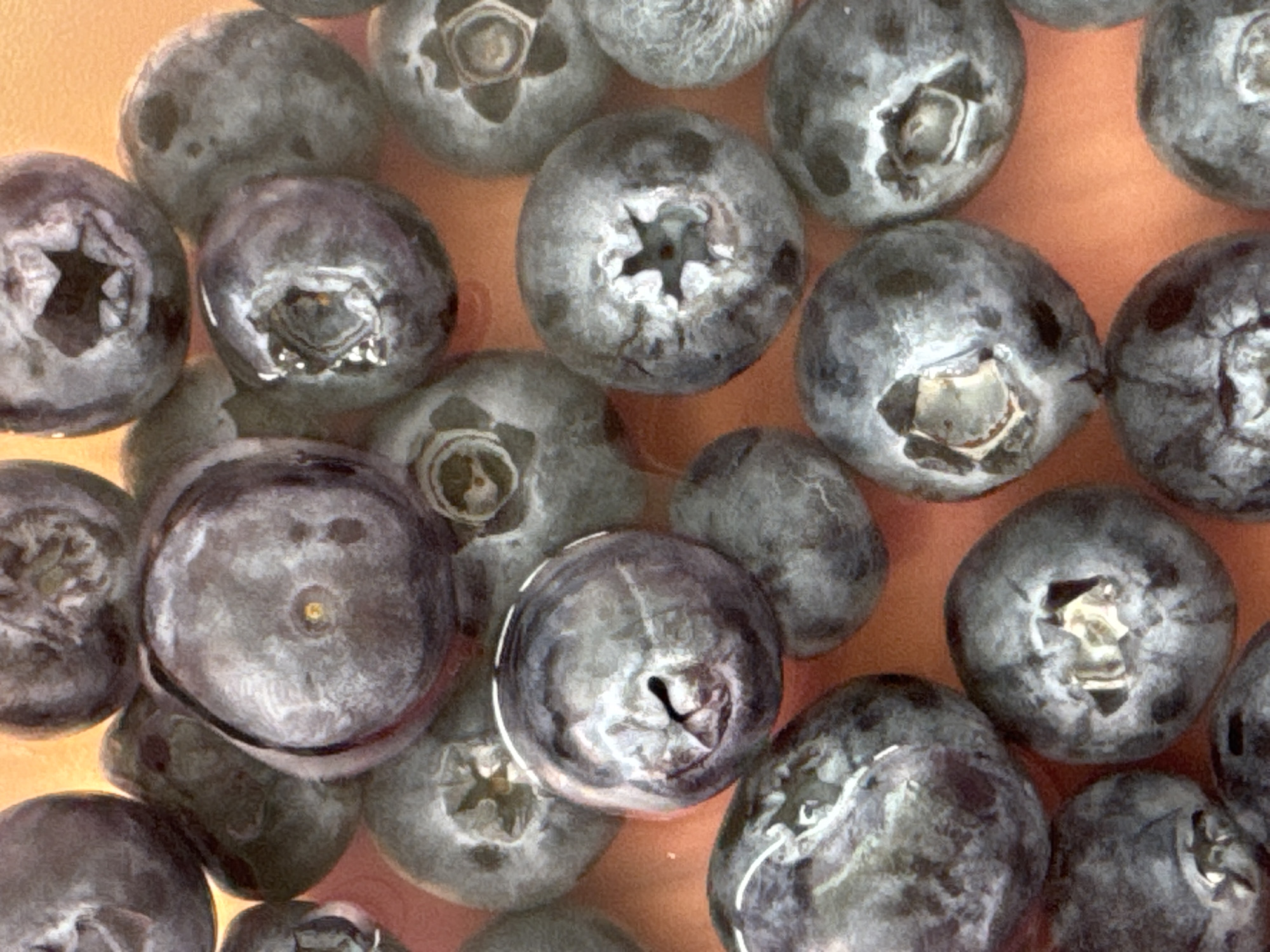 This is a close-up, overhead shot of a collection of fresh blueberries, filling the entire frame. The berries are various shades of deep blue and purple, with some appearing slightly lighter due to the lighting and the natural bloom on their skins. The smooth, rounded surfaces of the blueberries are textured with a matte finish, and many of them show the characteristic star-shaped calyx remnants at their tops. Some berries are partially obscured by others, creating a sense of depth and abundance. The background is a soft, warm orange-brown color, likely the surface they are resting on or a shallow liquid they are submerged in, which allows the vibrant blue of the blueberries to stand out. The lighting highlights the subtle curves and indentations of each berry, emphasizing their natural forms. There is no text visible in the image. The scene appears to be a simple, still life focusing solely on the fruit. The context provided suggests the location is Moscow, Russia, but this is not visually indicated within the image itself.