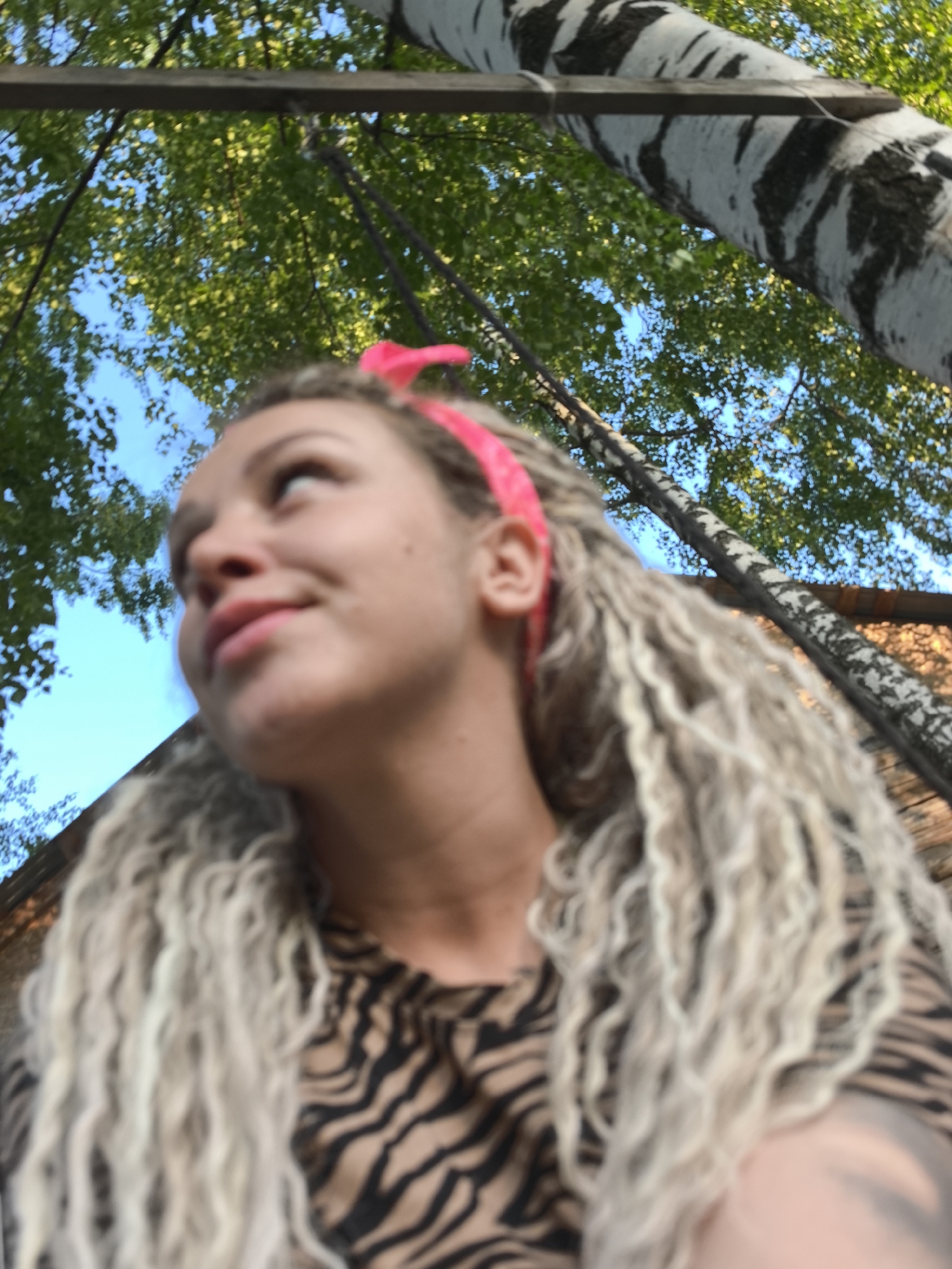 A low-angle shot depicts a woman with blonde curly hair and a pink headband. She is wearing a black and brown patterned shirt. Her gaze is directed upwards and to the left. Above her, there are green tree leaves and a blue sky. A black and white birch tree trunk is visible in the upper right corner of the image. A wooden beam also appears to be present among the trees. The location may be a park or wooded area based on the presence of trees. Based on the prompt, this scene is located in the city of Noviy, Russia.
