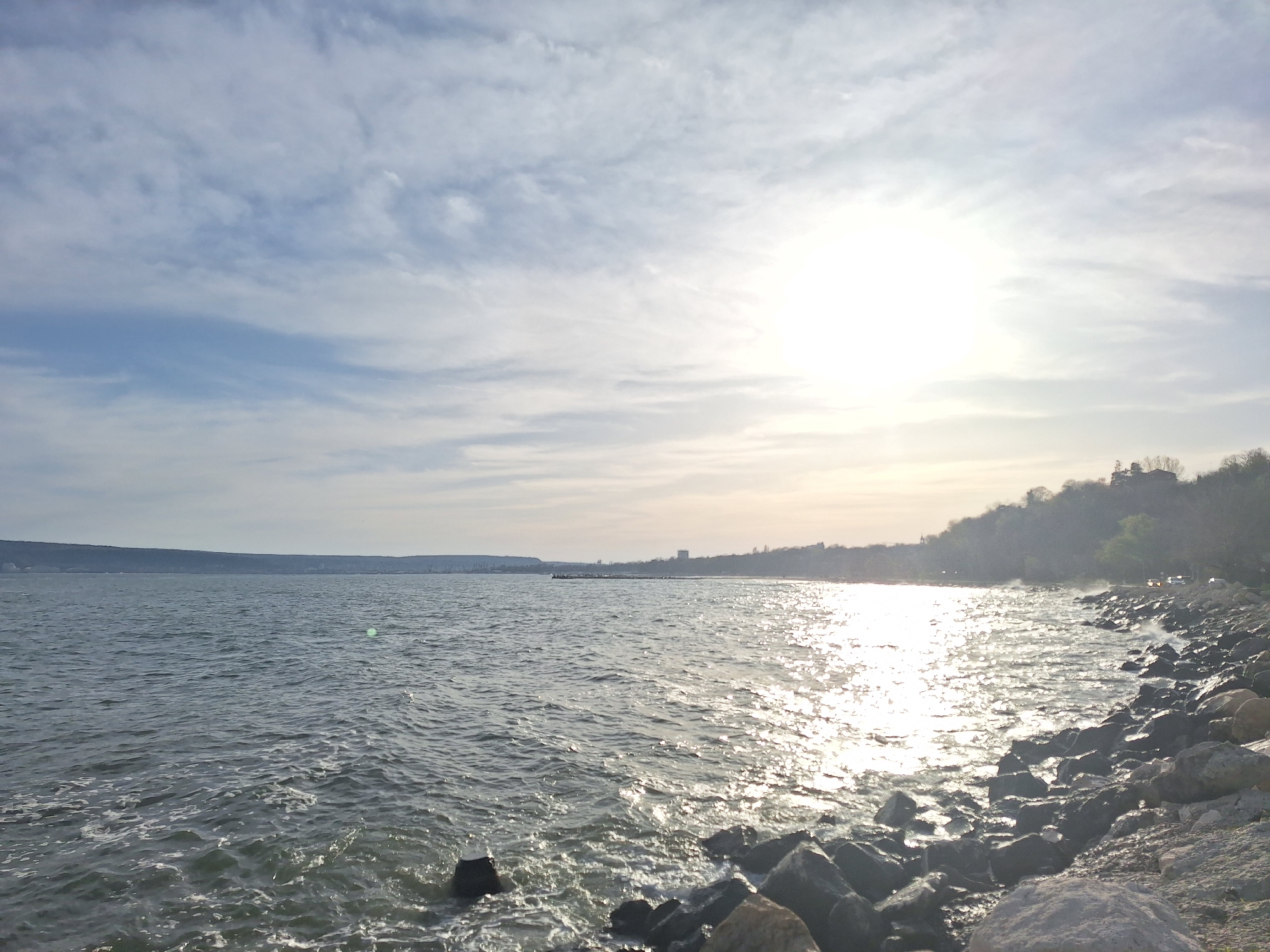 The image depicts a seascape near Varna, Bulgaria, characterized by a rocky shoreline on the right side of the frame and the Black Sea extending to the left. The sea is moderately choppy, reflecting sunlight, creating a shimmering effect. The horizon is punctuated by a landmass in the distance, possibly marking the opposite side of the bay or the coastline stretching beyond Varna. The sky is bright with scattered clouds and the sun is high. The rocks on the shoreline are various sizes. A coastal road with limited visibility is present behind the trees. The image is likely taken in the early morning or late afternoon.