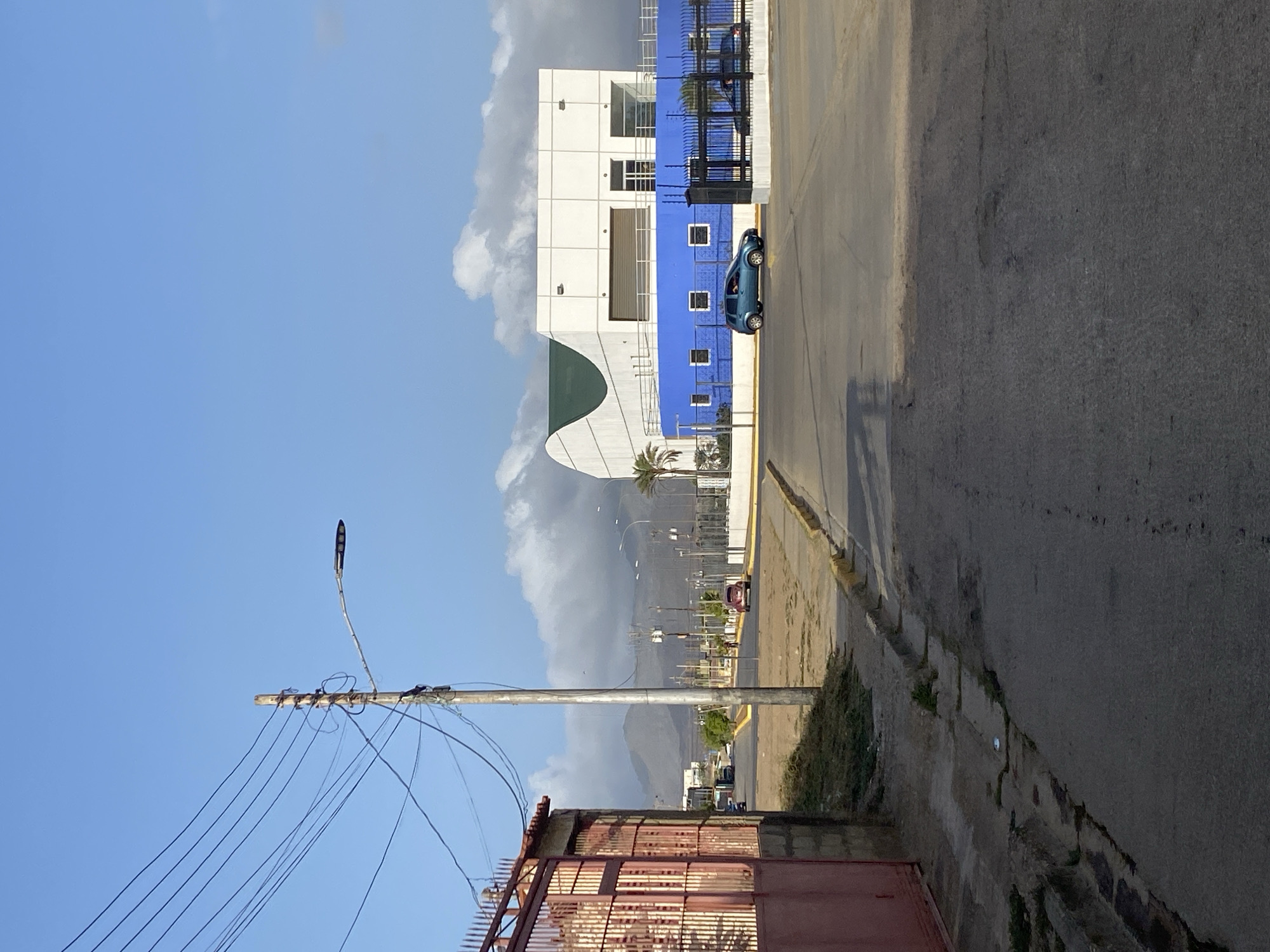 The image shows a street scene in Juan Griego, Venezuela. A white and blue building with a curved green awning is visible to the left of the road. A blue car is parked near the building. In the foreground, there is a rusted metal fence and a utility pole with numerous wires extending from it. The road stretches into the distance, where other buildings and cars are visible. A palm tree stands in the median of the road. Mountains are seen in the background, partially obscured by clouds. The sky is blue with scattered clouds. The scene is viewed from a slightly elevated position, possibly from a sidewalk or building. The angle of the photograph has been rotated 90 degrees.