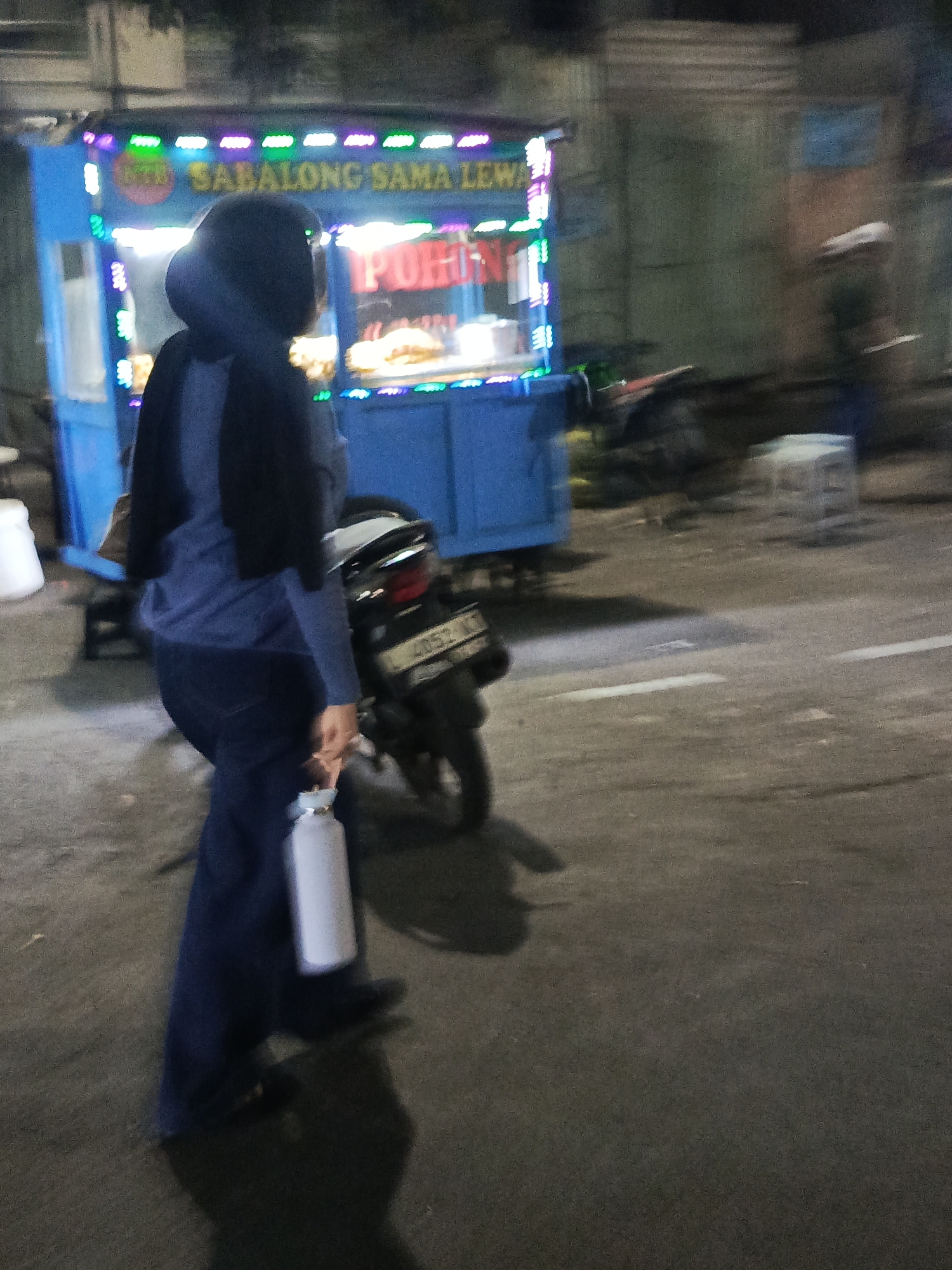 A person wearing a dark headscarf and a blue long-sleeved shirt is walking on a street at night. They are holding a white cylindrical object, possibly a water bottle, in their right hand. Behind the person is a food cart with blue and green lights, displaying the text "SABALONG SAMA LEW" and "POHN". A motorcycle is parked next to the food cart. The scene appears to be in a public area, likely a street or market, in Surabaya, Indonesia. The ground is asphalt, and there are streetlights contributing to the illumination. The image is slightly blurred due to motion.